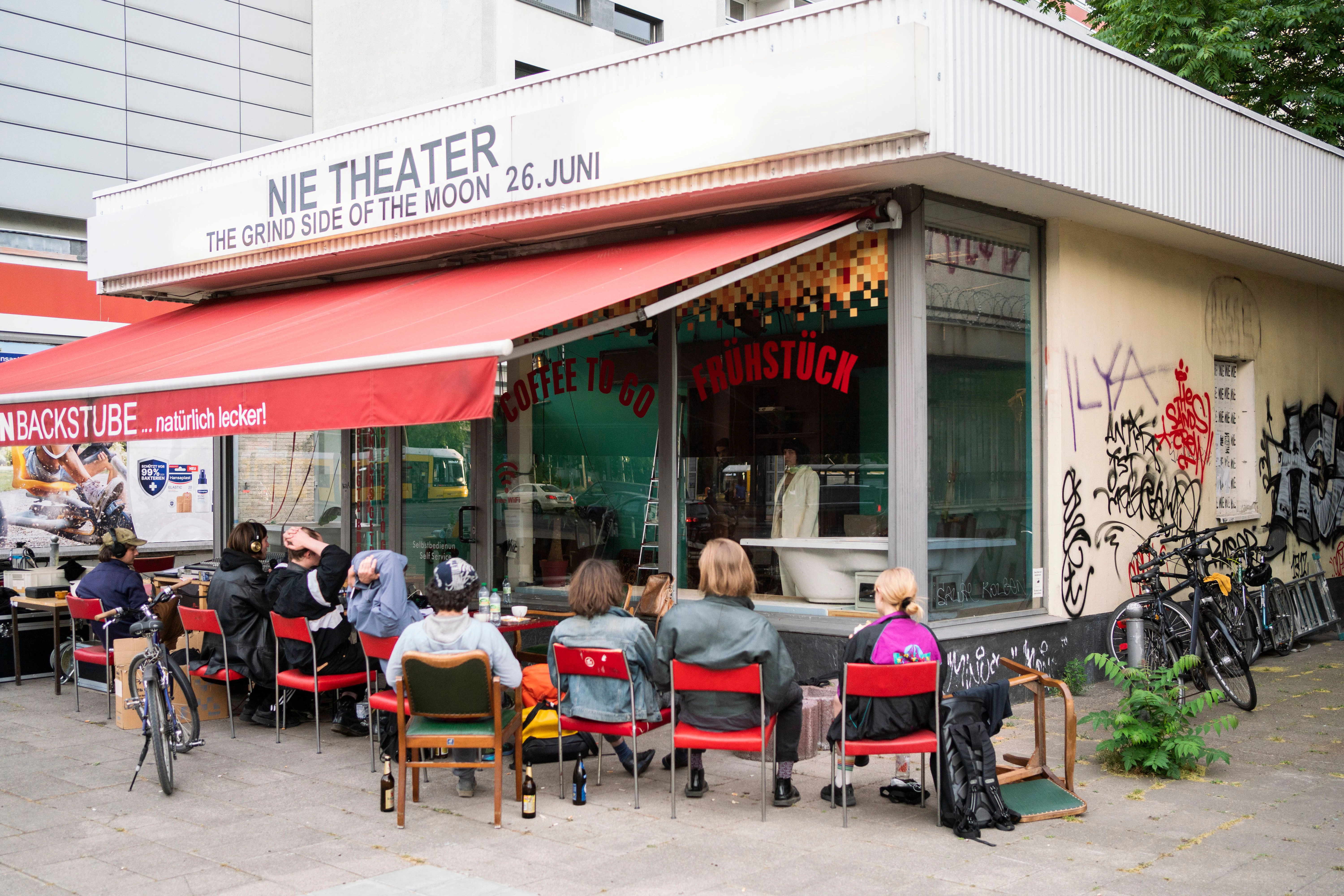 Menschen sitzen vor einer ehemaligen Bäckerei am Alexanderplatz, die in ein Theater umgewandelt wurde. Berlin, 2021.