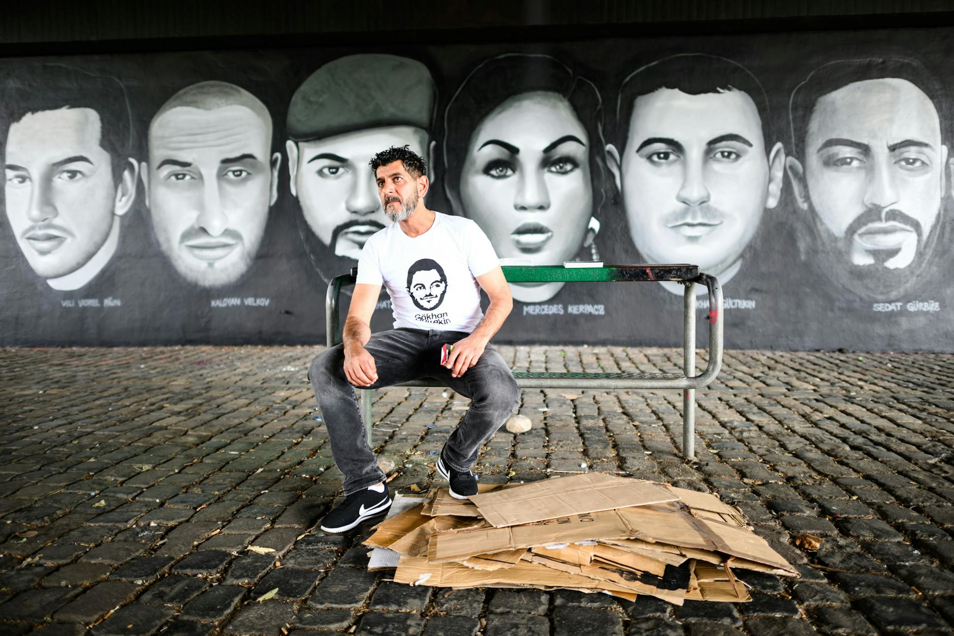 Cetin Gültekin, Bruder des Opfers Gökhan Gültekin, sitzt vor Porträts von Opfern des Anschlags von Hanau. Ein Gemälde unter der Frankfurter Friedensbrücke zeigt ihre Gesichter.