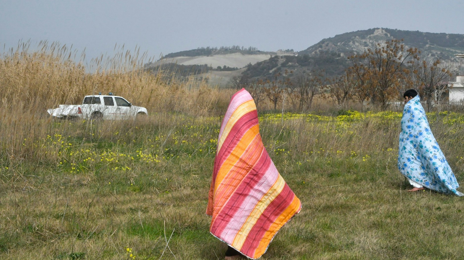 Zwei Überlebende entfernen sich, in bunte Decken gehüllt, vom Ufer des Mittelmeers.