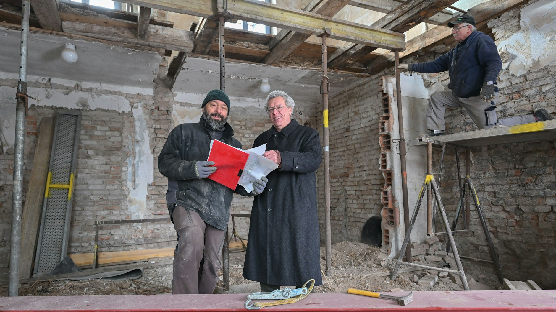 Bauunternehmer Marko von Cysewski (l.) und Pfarrer Thomas Berg stehen auf der Baustelle eines alten Hauses, wo einmal ein Hofladen entstehen soll. Auf dem Gerüst arbeitet Eberhard Netzker.