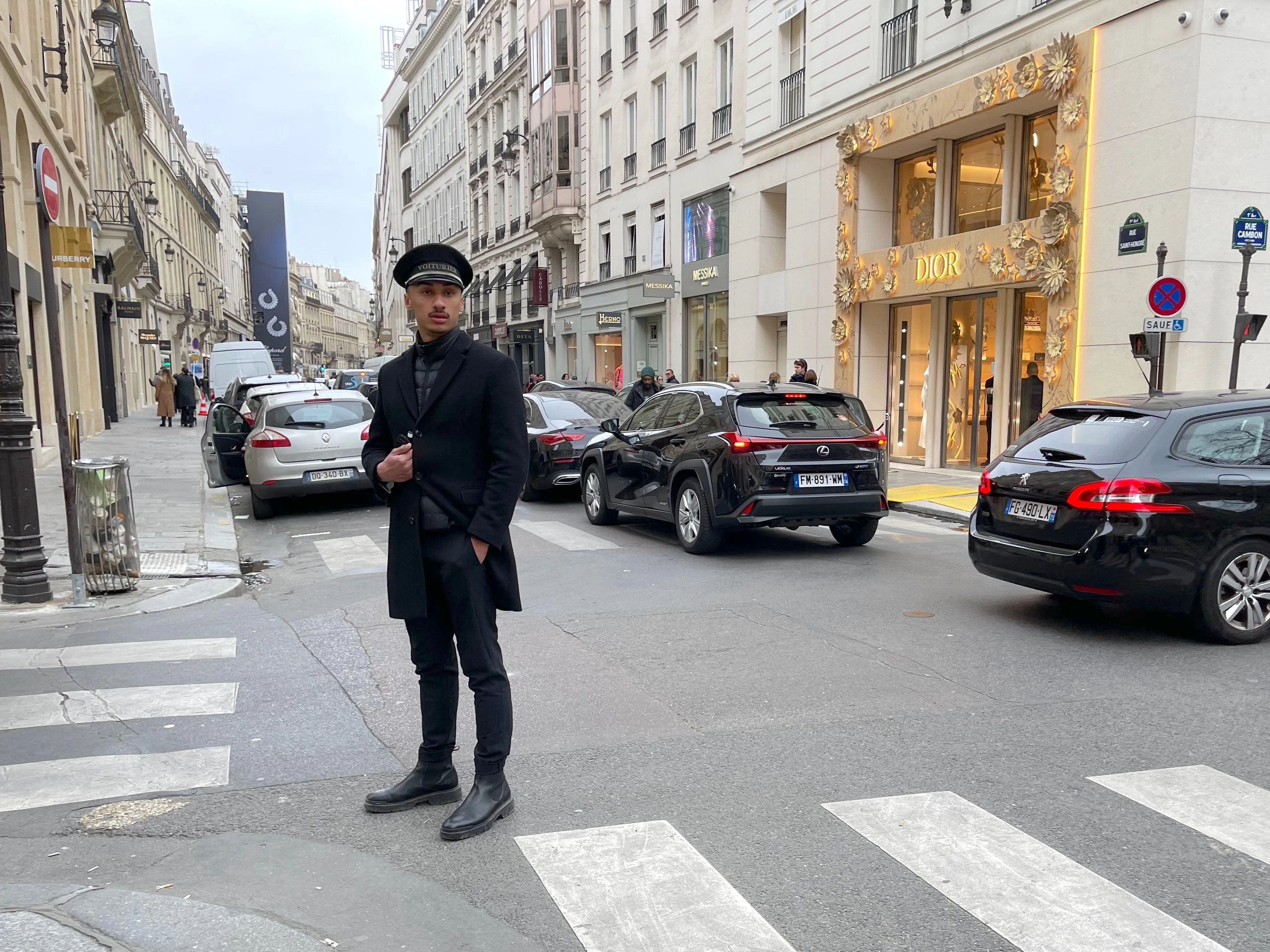 Der Herr hat das Wort „Voiturier“ an der Mütze stehen. So werden die Angestellten eines Luxushotels bezeichnet, die für das Einparken und Vorfahren der Gästewagen zuständig sind.