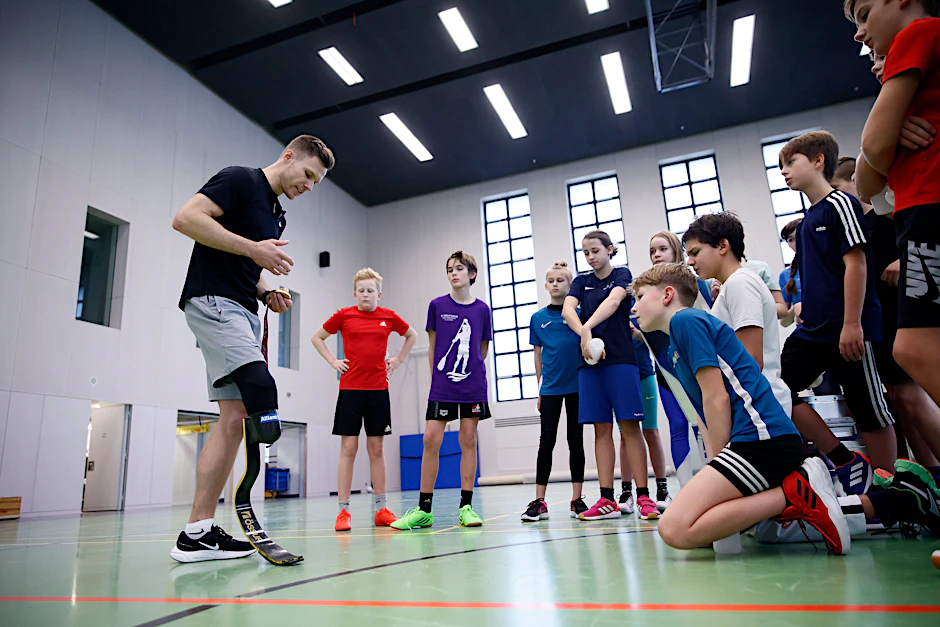 Weitspringer Markus Rehm spricht mit Schülern in einer Turnhalle bei dem Besuch der Sportschule im Olympiapark – Poelchau-Schule.
