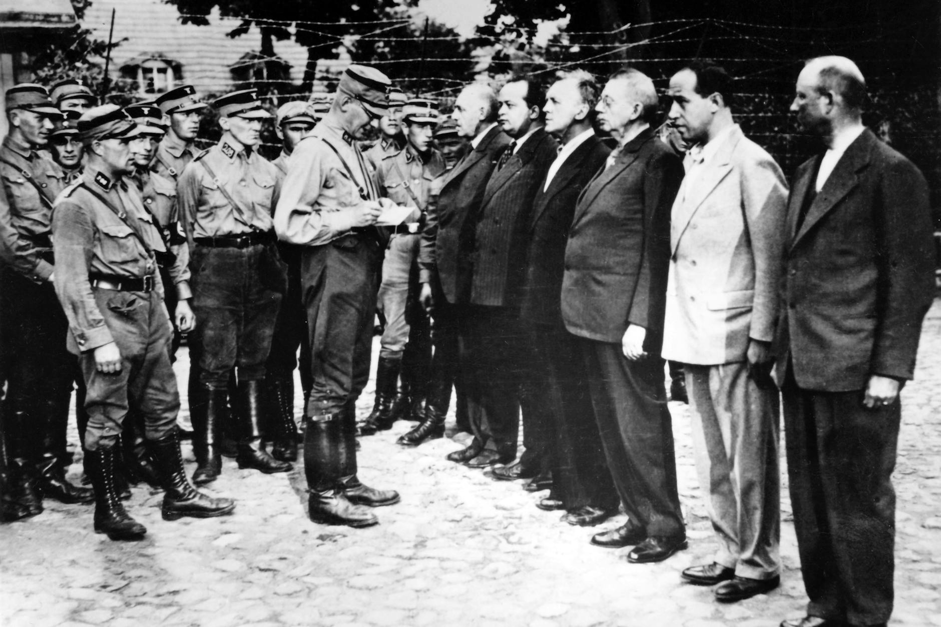 August 1933: Im Lagerhof des Konzentrationslagers Oranienburg stehen SA-Männer vor Parteifunktionären der SPD und Angehörigen des Rundfunks, v.l.n.r. Ernst Heilmann (SPD), Friedrich Ebert jun. (SPD), Alfred Braun (Reporter beim Berliner Rundfunk, Schauspieler, Regisseur), Heinrich Giesecke (Direktor der Reichs-Rundfunk-Gesellschaft), Hans Flesch (Intendant des Berliner Rundfunks), Kurt Magnus (Vorstandsvorsitzender der Reichs-Rundfunk-Gesellschaft)