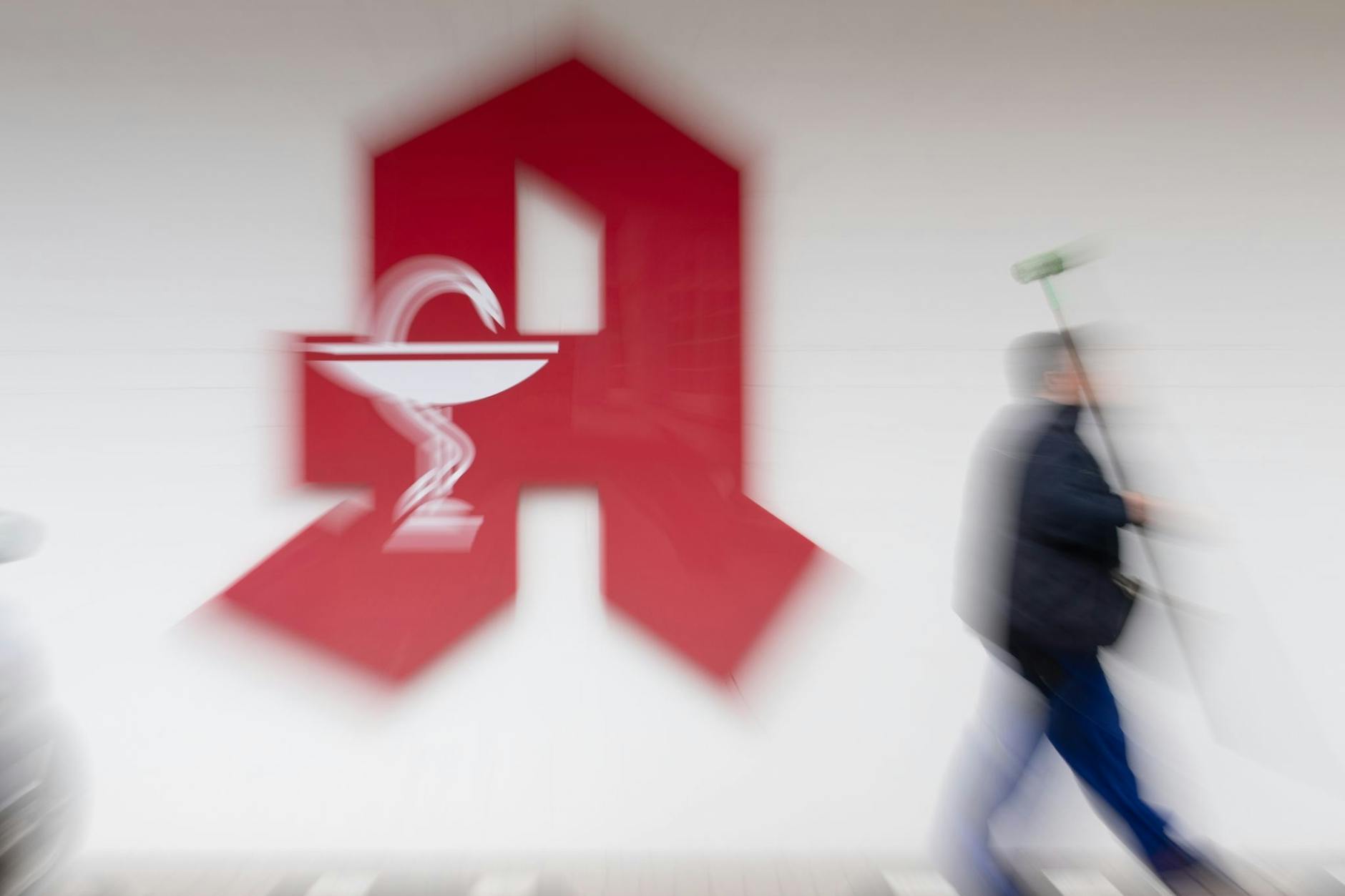 Ein Mann mit geschultertem Besen passiert ein Apotheken-Schild. 2022 gaben fast 400 Apotheken auf.