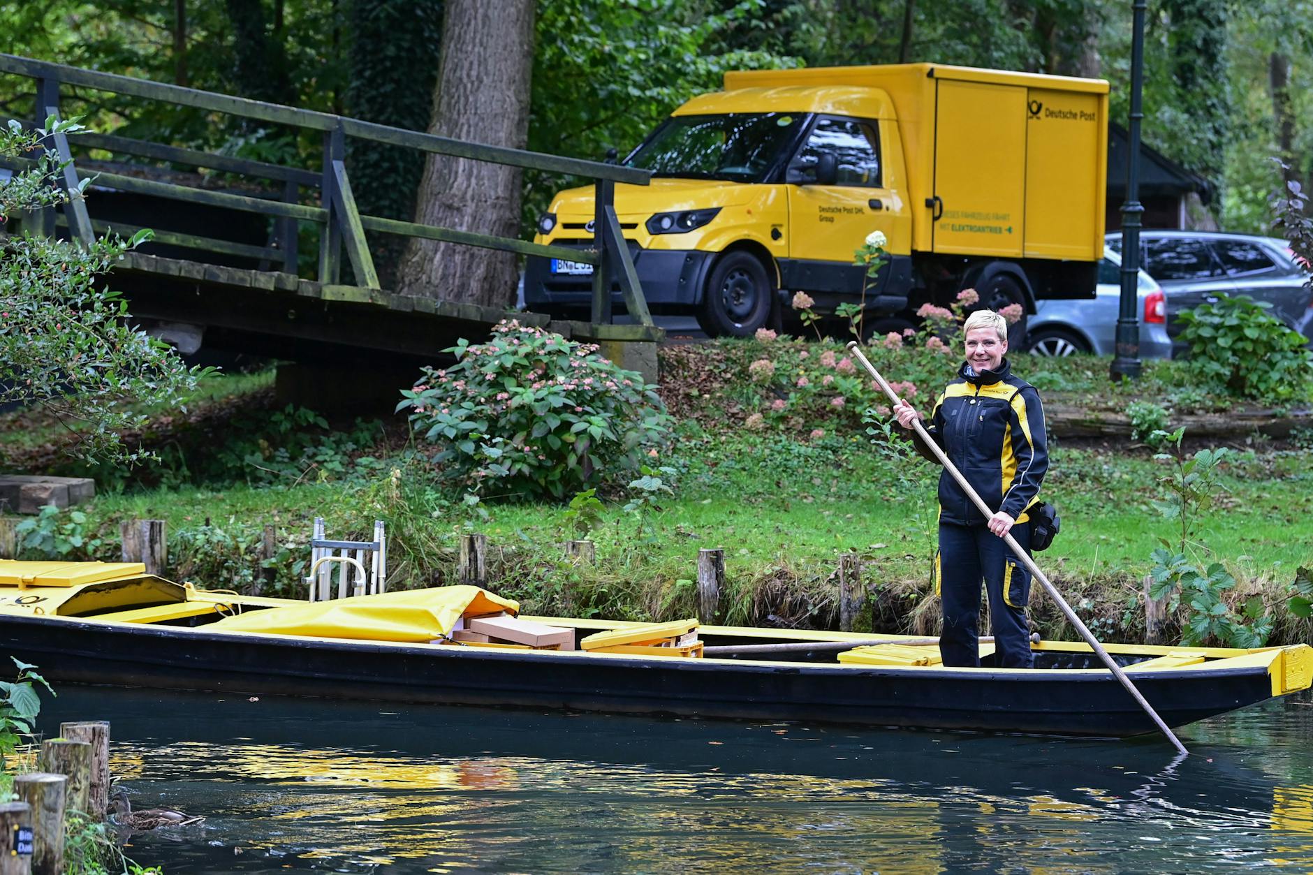 Andrea Bunar, Zustellerin der Deutschen Post, ist in ihrem gelben Postkahn unterwegs, während am Ufer ihr neues Elektrofahrzeug steht.