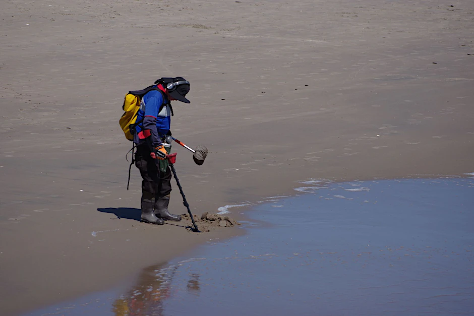 George Pessard mit einem Metalldetektor, Gummistiefeln, einem Sieb und zwei Plastikbehältern am Strand von Coney Island.  
