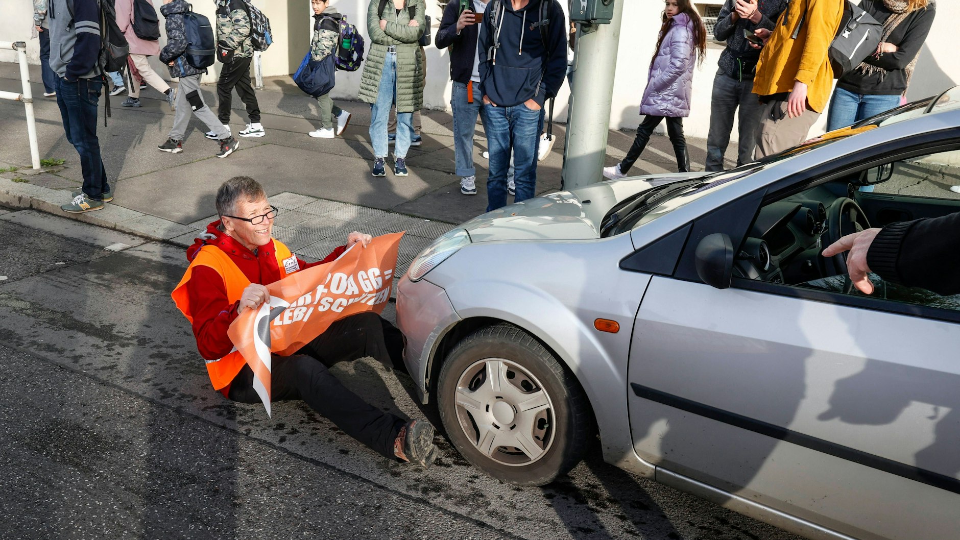 Ein Klima-Kleber setzt sich in der Kniprodestraße direkt vor einem Auto, blockiert so die Weiterfahrt des Wagens.