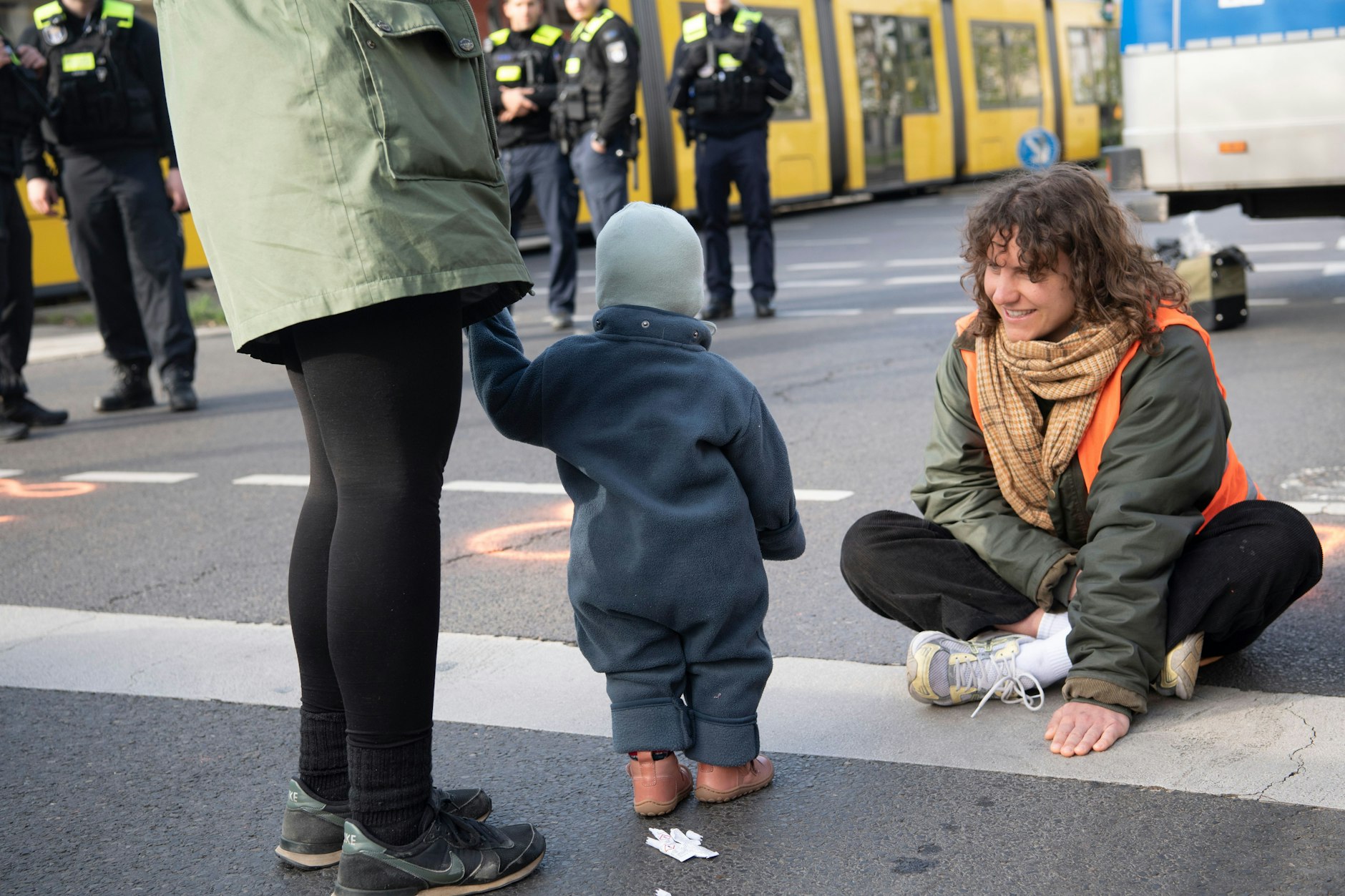 Eine Frau mit einem Kind steht bei einer Blockade der Gruppierung Letzte Generation auf einer Kreuzung an der Landsberger Allee vor einer Aktivistin, die die Straße blockiert.
