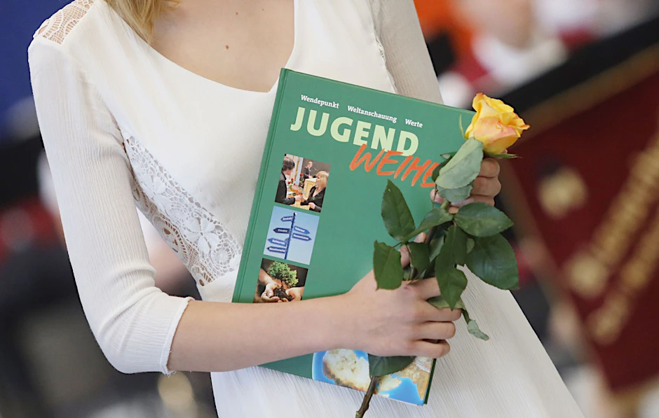 ARCHIV - Eine Teilnehmerin einer Jugendweihefeier hält eine Rose und ein Buch in der Hand.  