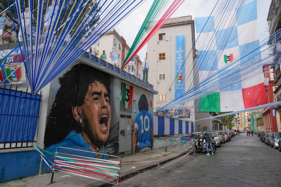 Ein riesiges Wandgemälde von Diego Armando Maradona und Fahnen vom SSC Neapel zieren eine Straße in Neapel.  