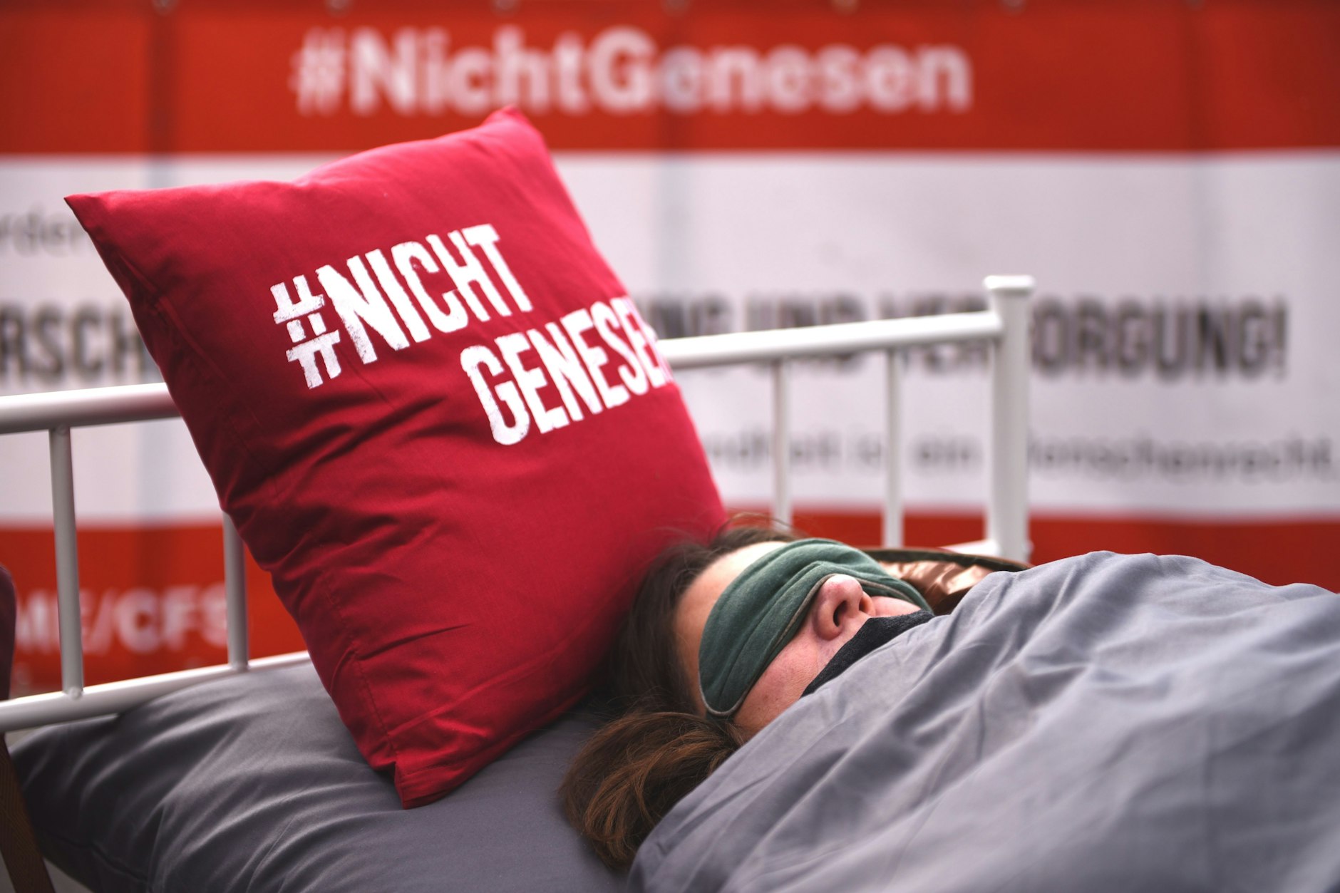 Eine Frau liegt mit Augenbinde im Bett. Sie demonstriert vor dem Wissenschaftsministerium in Berlin für mehr Fatigue-Forschung.