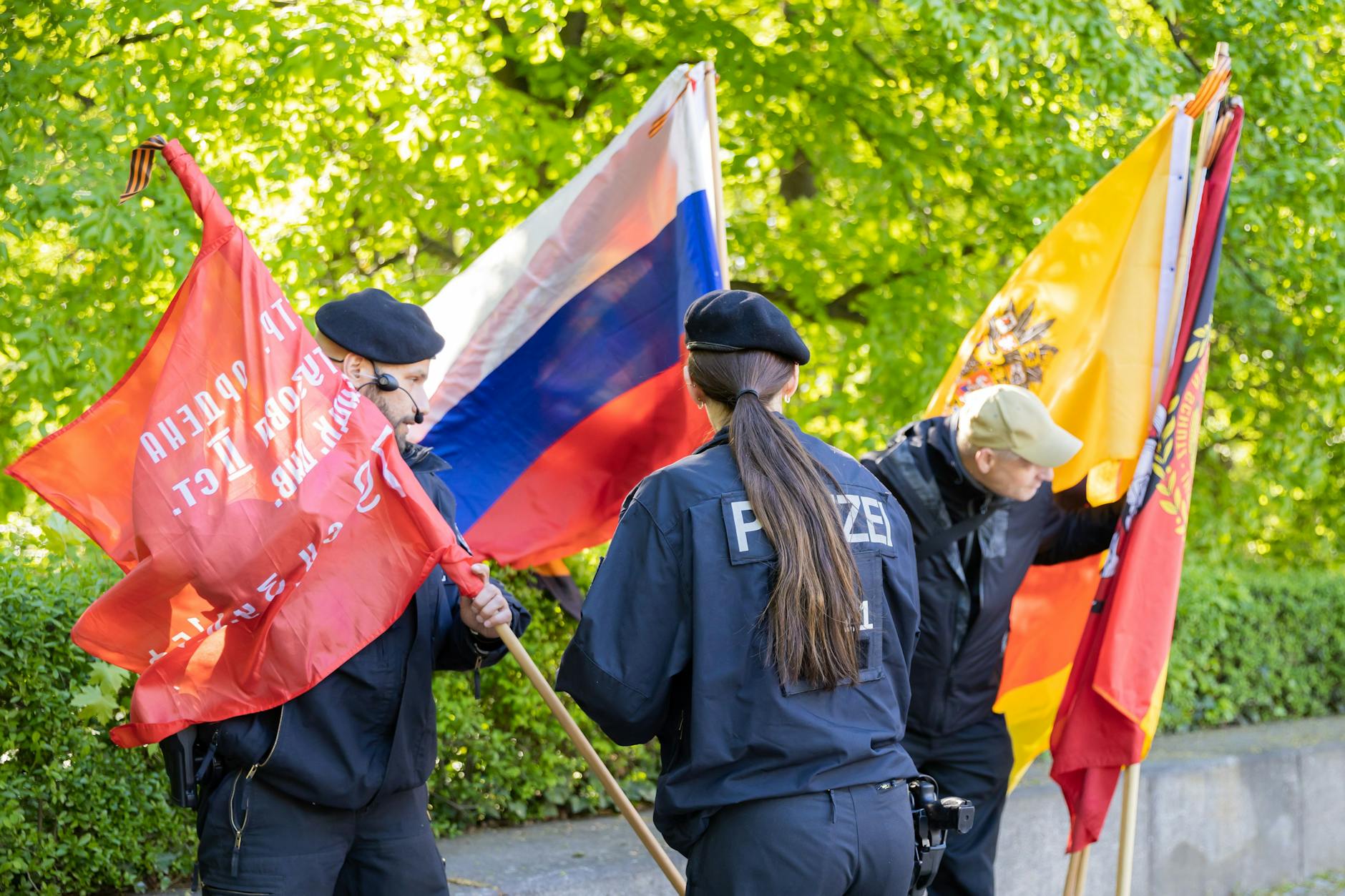 Polizisten kontrollieren am Sowjetischen Ehrenmal im Treptower Park die Flaggen eines Besuchers, wobei eine russische Flagge zu sehen ist.