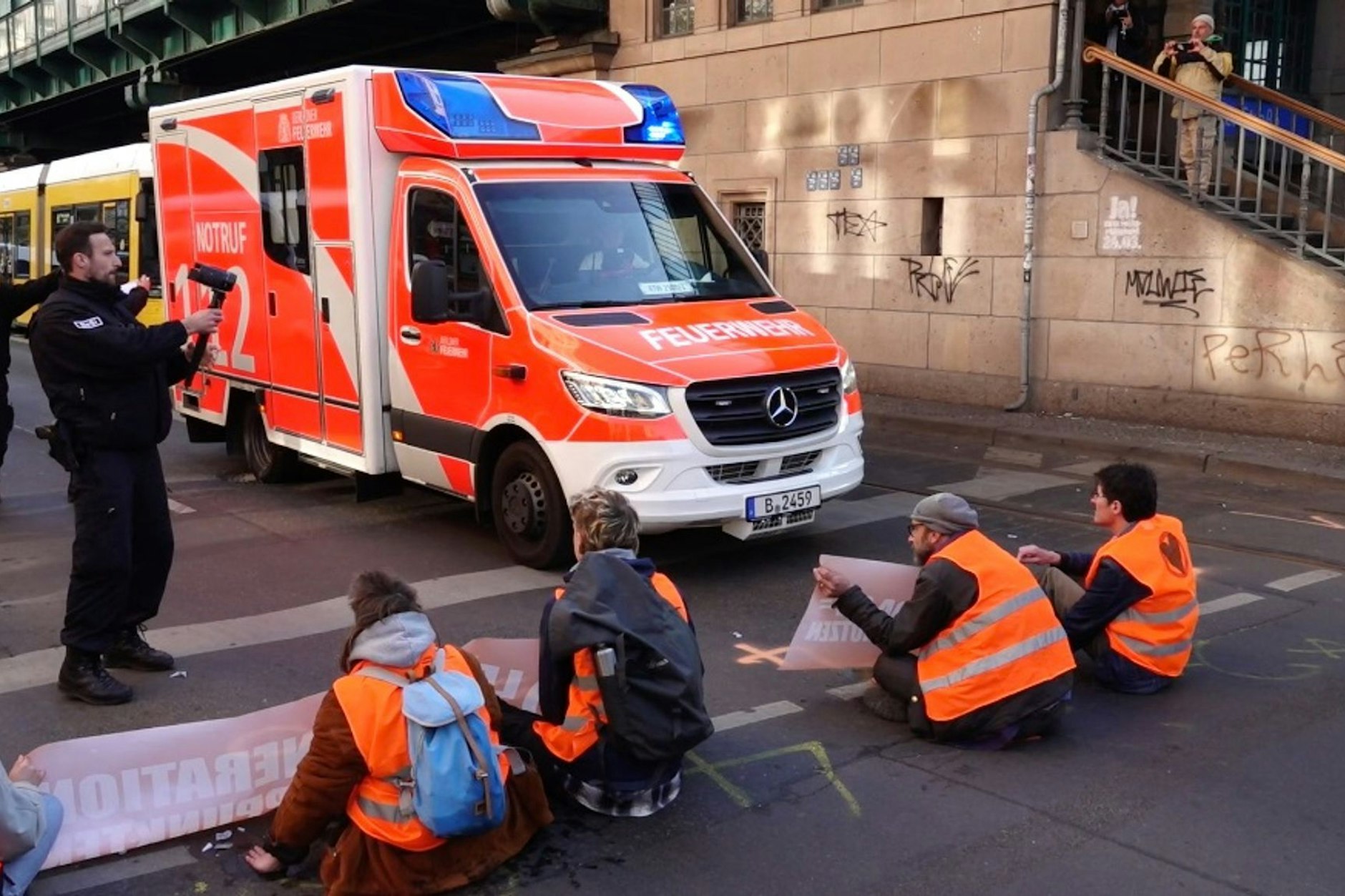 Aktivisten der Gruppe Letzte Generation blockieren die Fahrbahn an der Schönhauser Allee, Ecke Eberswalder Str., während ein Rettungswagen zum Einsatz unterwegs ist und daran vorbeifährt.