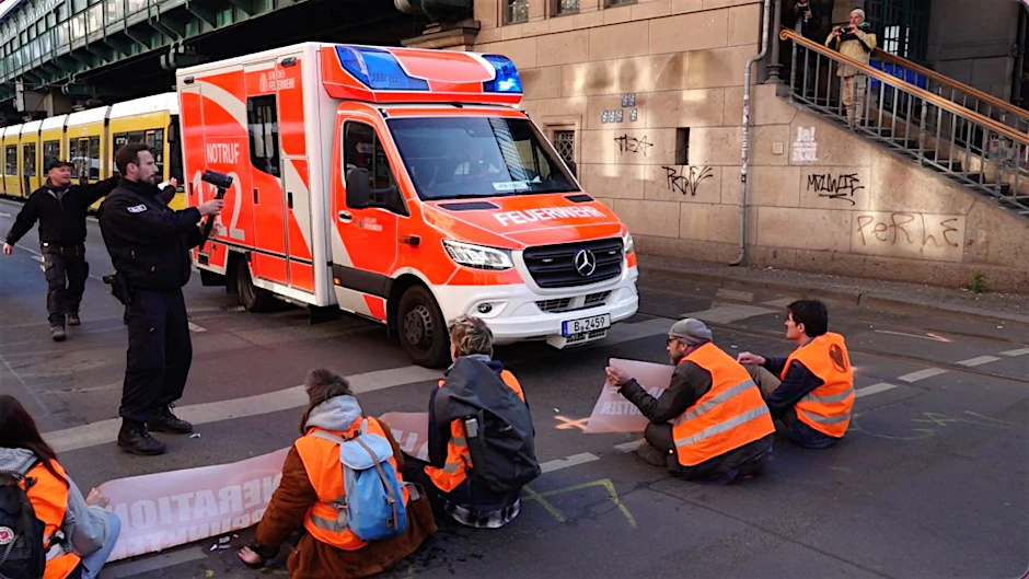Aktivisten der Gruppe Letzte Generation blockieren die Fahrbahn an der Schönhauser Allee, Ecke Eberswalder Str., während ein Rettungswagen zum Einsatz unterwegs ist und daran vorbeifährt.
