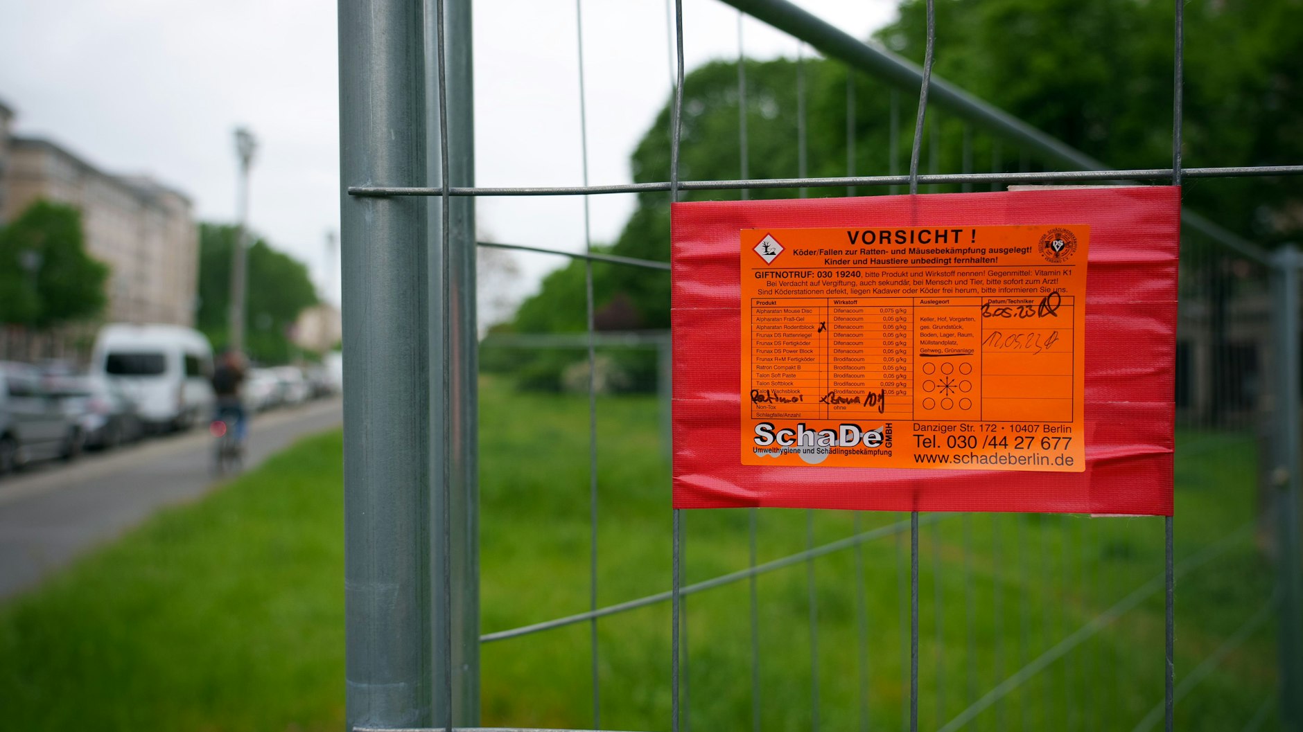 Auf der Wiese neben der Karl-Marx-Allee warnt an einem Metallzaun ein Schild vor einer Köder-Falle für die Rattenbekämpfung. 