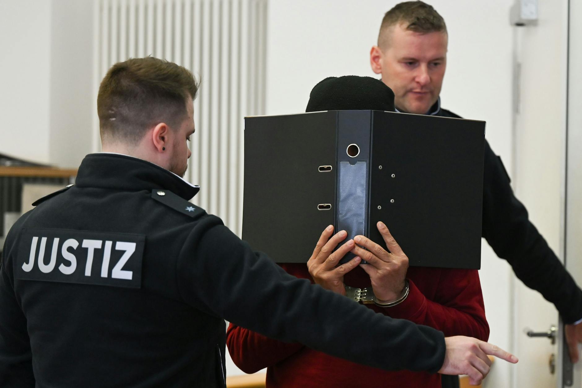 Justizbeamte führen den 47-jährigen Angeklagten (M.), der Handschellen trägt und sein Gesicht hinter einem Aktenordner versteckt, zum Prozessauftakt in den Gerichtssaal des Landgerichts.