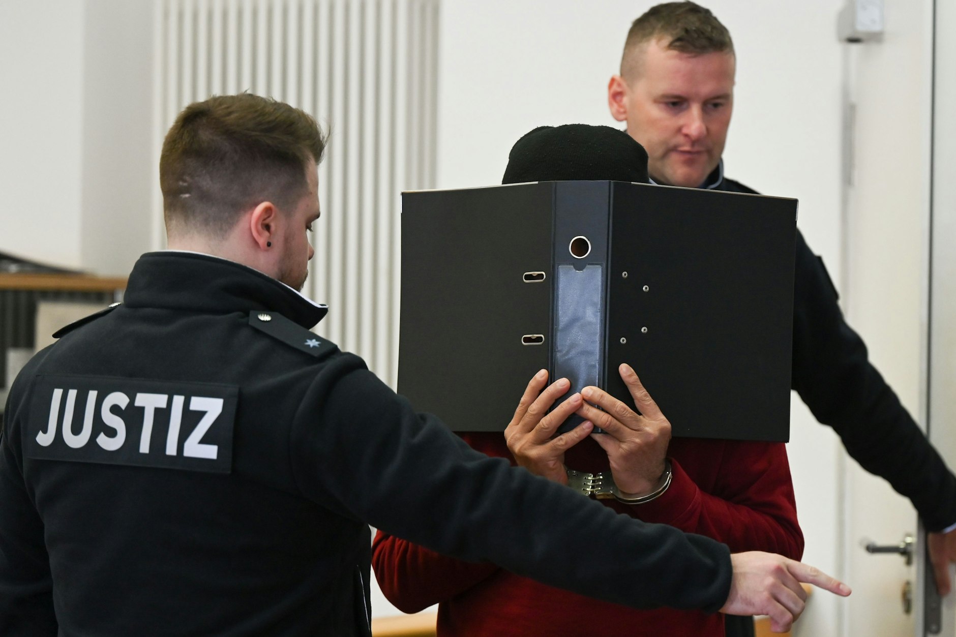 Justizbeamte führen den 47-jährigen Angeklagten (M.), der Handschellen trägt und sein Gesicht hinter einem Aktenordner versteckt, zum Prozessauftakt in den Gerichtssaal des Landgerichts.