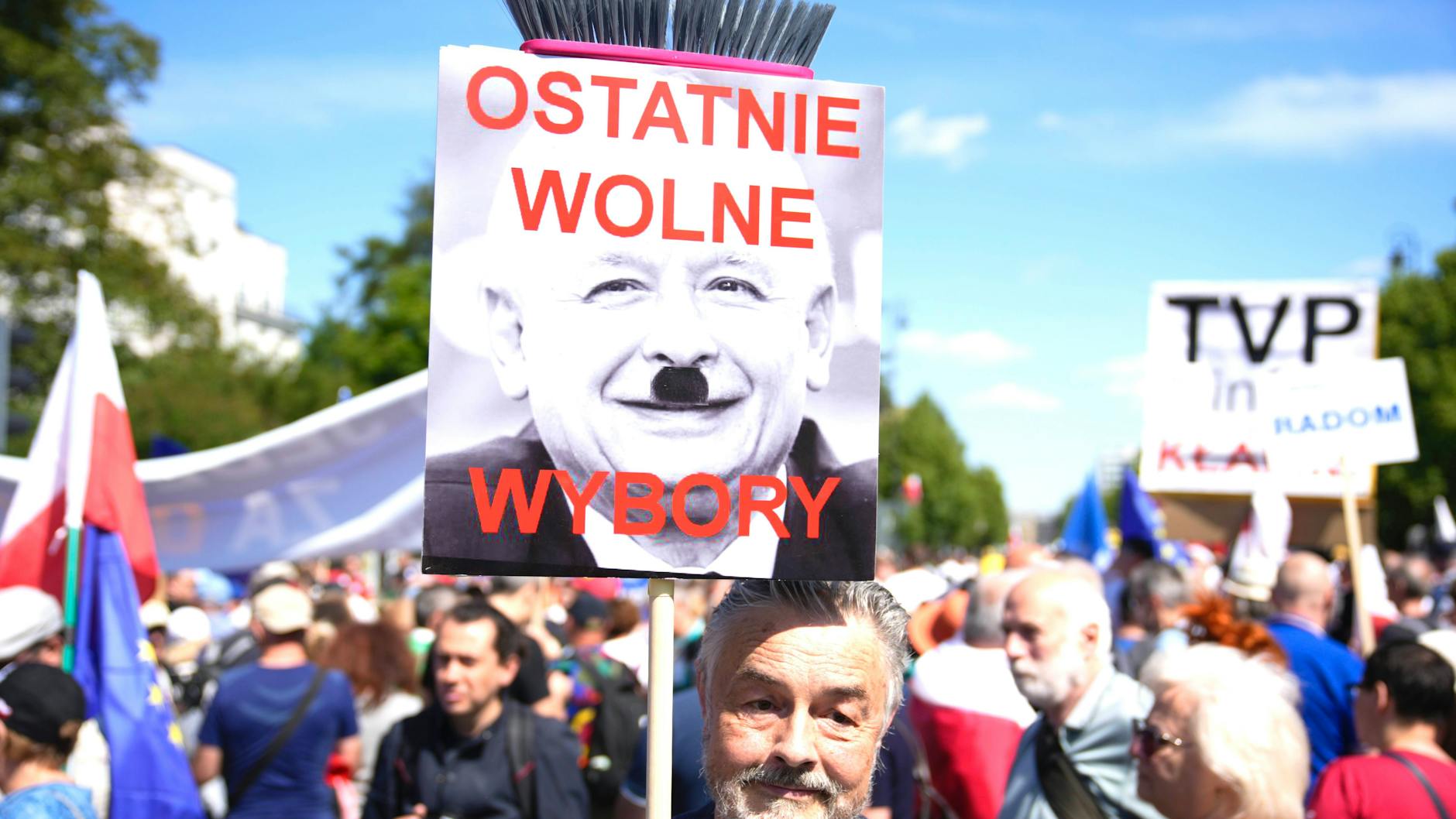 Ein Mann trägt ein Schild, an einem Besen befestigt. Darauf steht „Die letzten freien Wahlen“. Es zeigt Jaroslaw Kaczynski, graue Eminenz der PiS-Partei, mit Hitlerbärtchen.