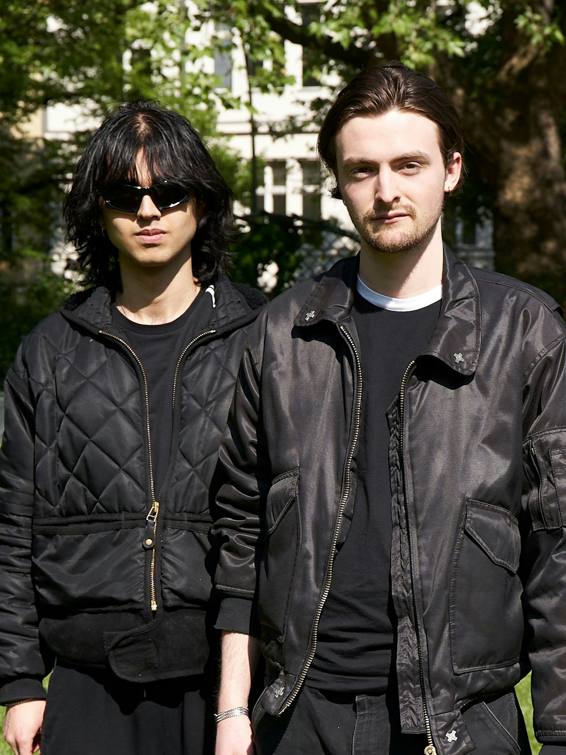 Elias trägt die „Bat-Rectangle“-Sonnenbrille von Balenciaga, eine gesteppte Jacke von 032c und ein T-Shirt von Adidas. Oscar trägt eine Bomberjacke mit Applikationen und abgeschnittenem Bund, die mal Elias gehört hat. Darunter hat Oscar ein schwarzes T-Shirt an, das ebenfalls von Adidas ist.