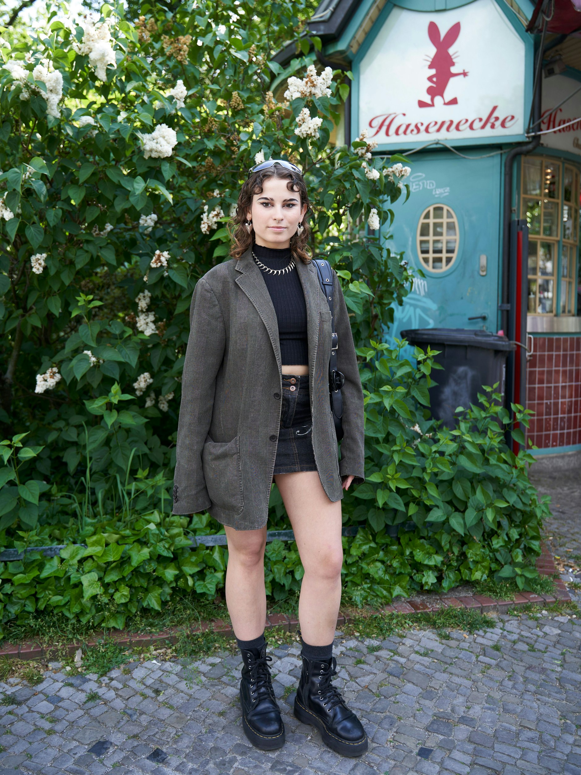 Alles Vintage, sagt die Studentin der Sozialwissenschaften über ihre Kleidung. Zu Cropped-Top, Jeans-Mini und Herrensakko trägt sie Dr. Martens mit Plateausohle. Die Statement-Herzohrringe hat sie von einem Spanienurlaub mitgebracht. Hinter Mara sieht man übrigens den legendären Mini-Imbiss „Hasenecke“ an der Ecke Kantstraße. Wird Maras Weg später noch ins Schwarze Café führen?