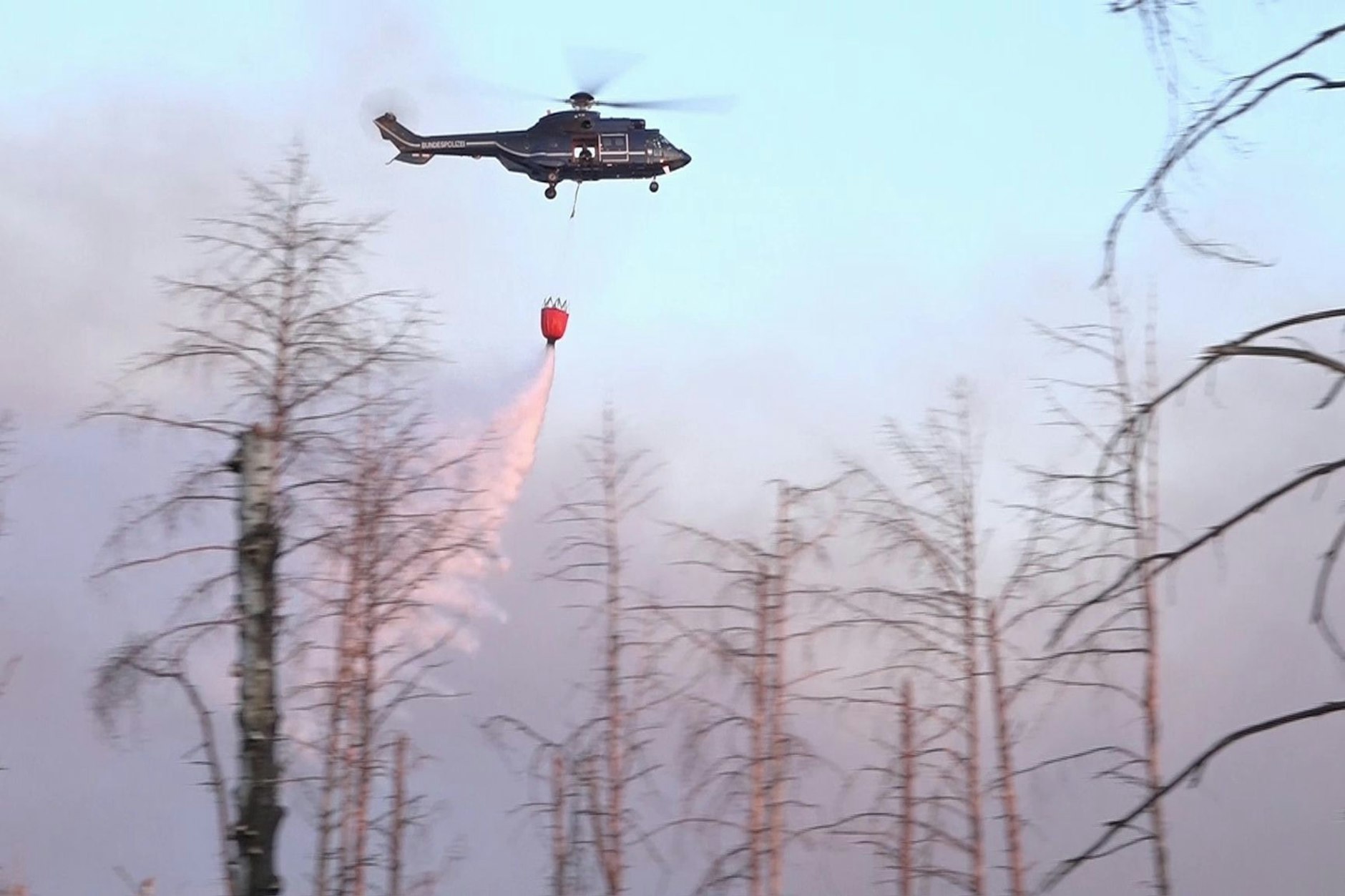 Ein Löschhubschrauber wirft Wasser über einem Waldbrand in einem munitionsbelasteten Gebiet ab.