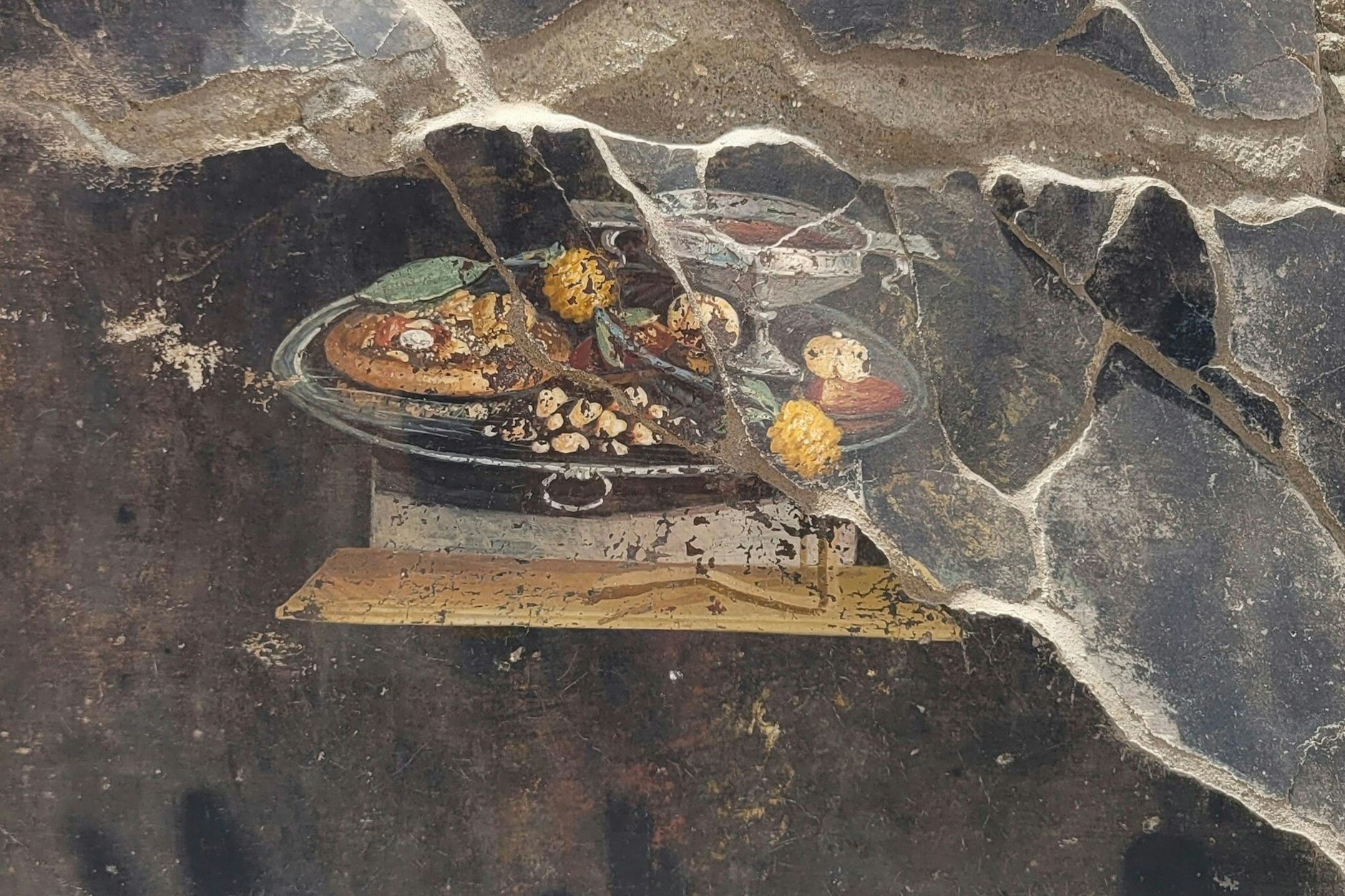 Die Wand eines Hauses in Pompeji mit einem Fresko, das einen Tisch mit Speisen darstellt, darunter auch eine Art Pizza.