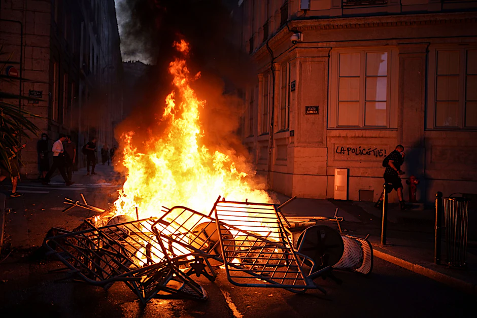 Ein Mann geht in Lyon neben einer brennenden Barrikade an einer Mauer vorbei, auf der "Die Polizei tötet" steht. 