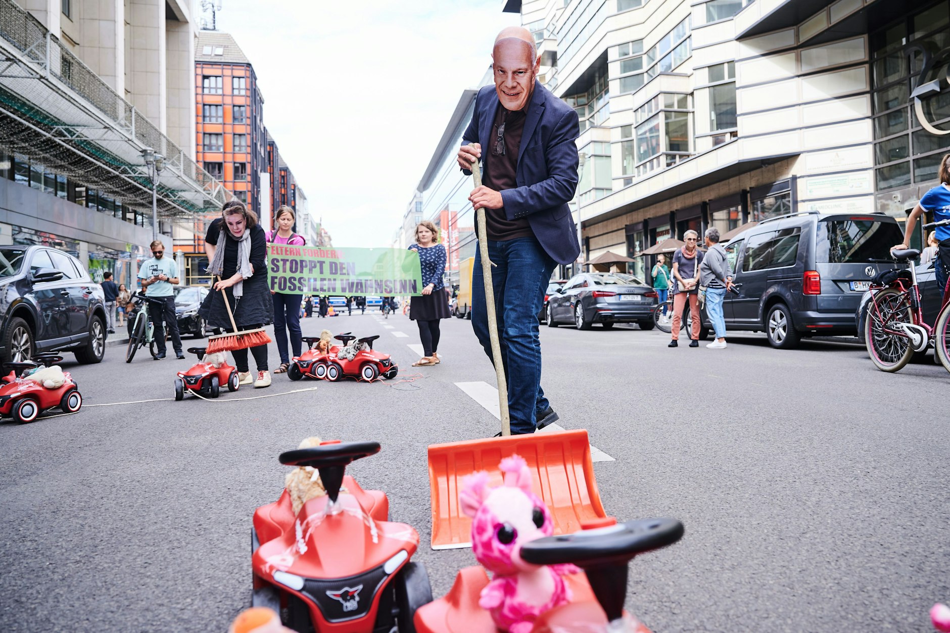 Ein Demonstrant mit einer «Kai-Wegner-Maske» kehrt symbolisch auf der Friedrichstraße die Bobby-Cars aus dem Weg. Die Elterngruppe "Eltern gegen die Fossilindustrie" protestiert gegen die Öffnung der Straße für den Autoverkehr.