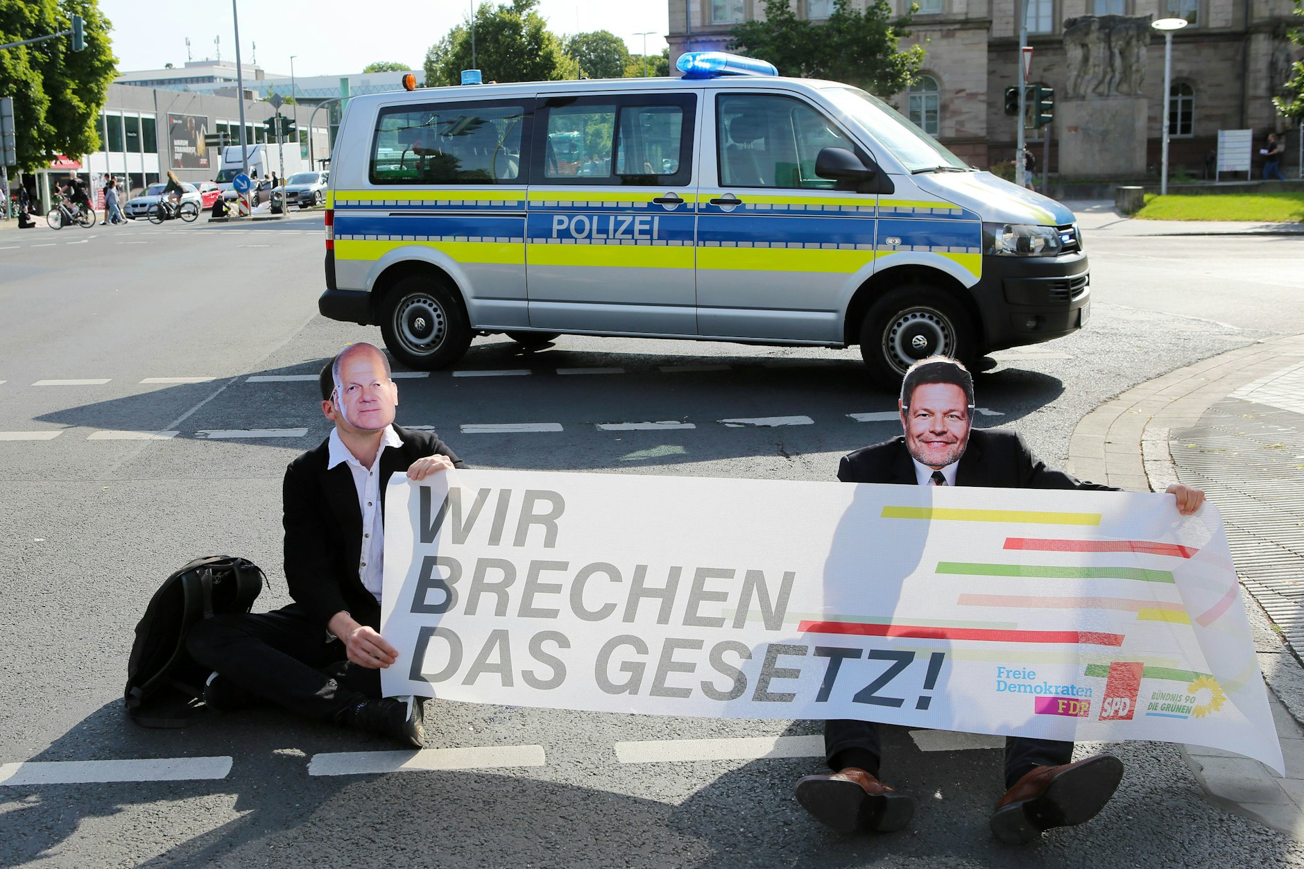 Göttingen: Klimaaktivisten der Gruppe Letzte Generation sitzen mit Masken von Bundeskanzler Scholz (l.) und Wirtschaftsminister Habeck bei einer Sitzblockade auf einer Kreuzung vor einem Polizeiauto.