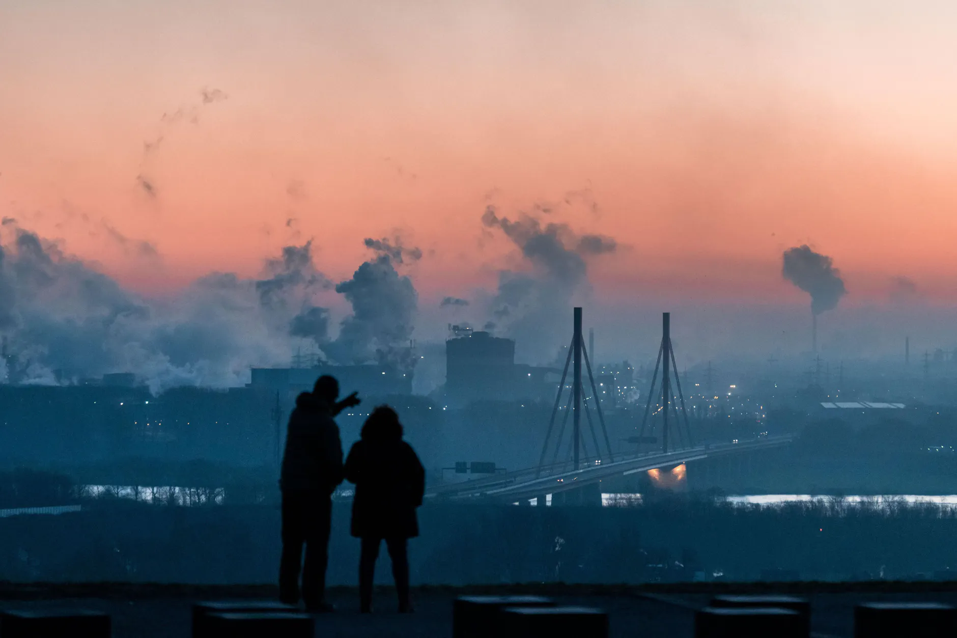 Zwei Menschen stehen bei Sonnenaufgang auf der Halde Rheinpreußen in Moers und gucken in Richtung des Industriegebiet von Thyssen-Krupp.