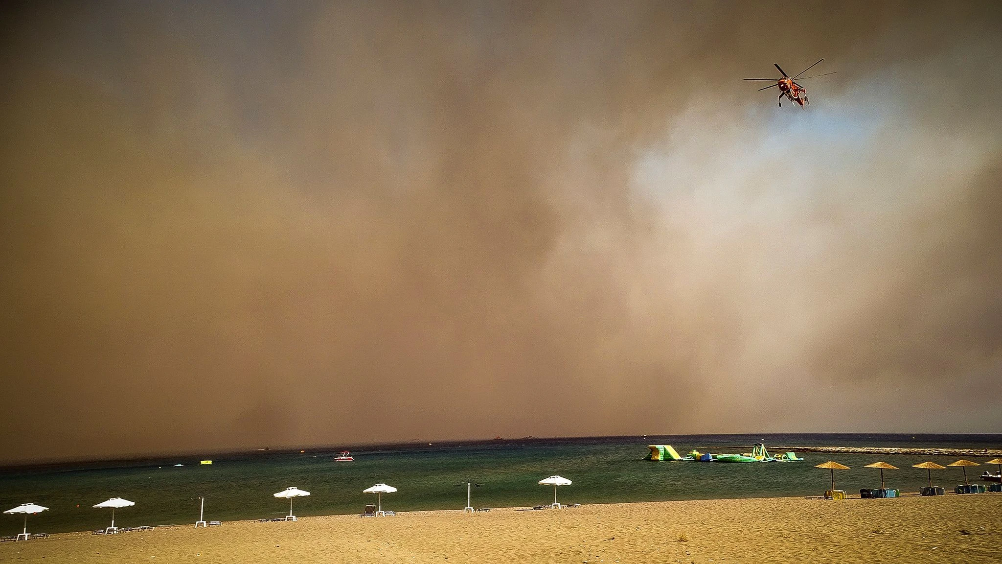 Ein Löschhubschrauber fliegt über einen Strand hinweg – und schwarzer Rauch verdunkelt das Badeparadies von Rhodos.