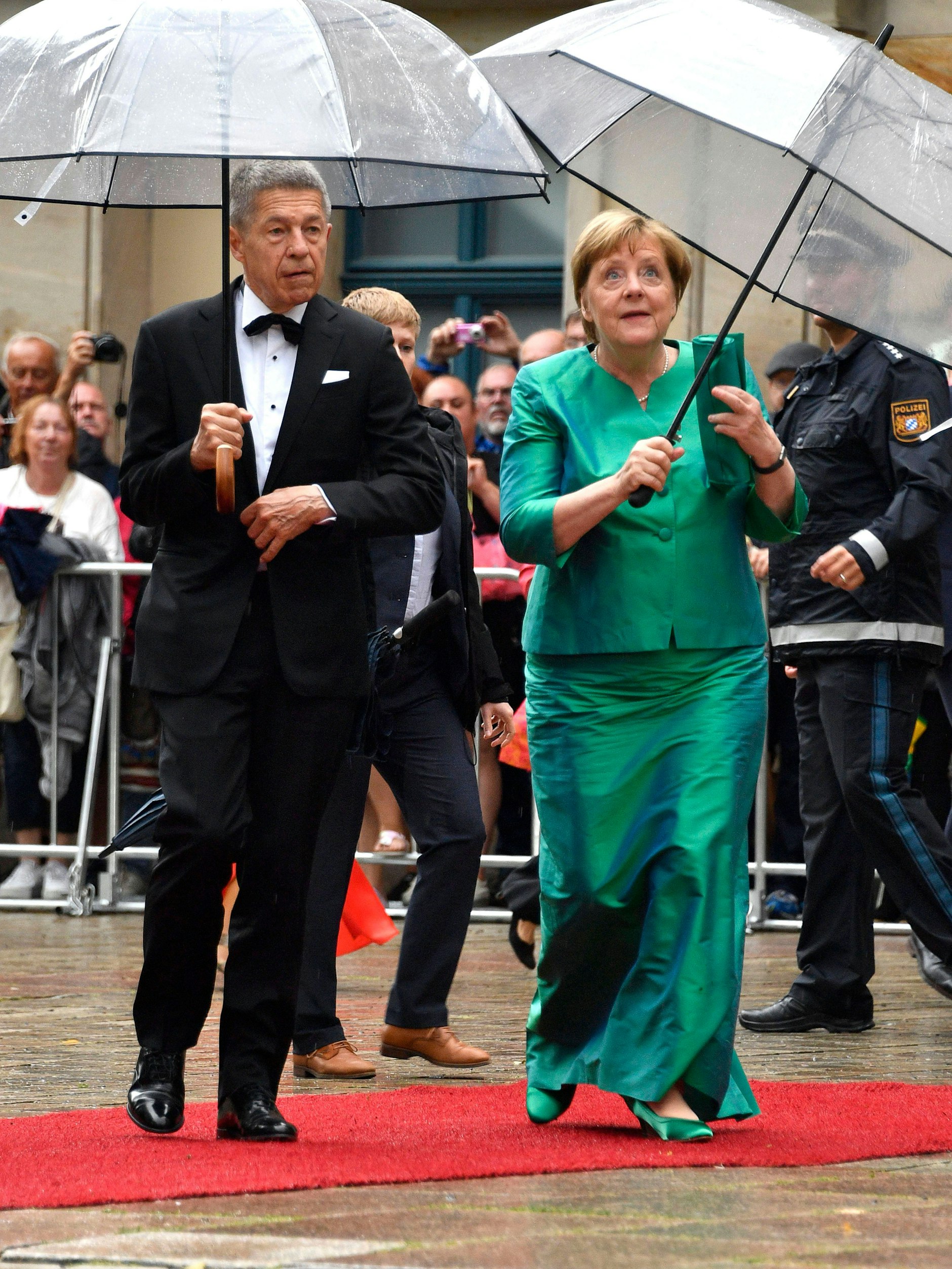 <strong>TOP:</strong> Kein Sinn für Progressivität, kaum Willen zur Veränderung, die Strategie des Aussitzens und Abwartens: Was Merkels Politik zur Farce gemacht hat, kommt ihr bei der Garderobenwahl zugute. Weil sie die stoische Unbeirrbarkeit vor größeren Peinlichkeiten bewahrt. So wie bei der Eröffnung der Richard-Wagner-Festspiele in Bayreuth, zu der sich Ex-Bundeskanzlerin <strong>Angela Merkel</strong> für ein grün-bläulich schimmerndes Komplet aus bodenlangem Rock und kragenlosem Jäckchen entschied. Ein aufregender Look? Ganz und gar nicht. Aber einer, der eben nicht unangenehm auffällt. Und: Ein Outfit, das absolut Merkel ist. Die Frau hat einen wenig aufregenden, aber wirklich eigenen Stil entwickelt; die steife Jacke mit bezogenen Knöpfen und Dreiviertelärmeln – das ist Merkel, unverkennbar. Weniger gelungen: Der übergroße Käppchenkragen und die traurig herunterhängende Fliege von <strong>Joachim Sauer</strong>.