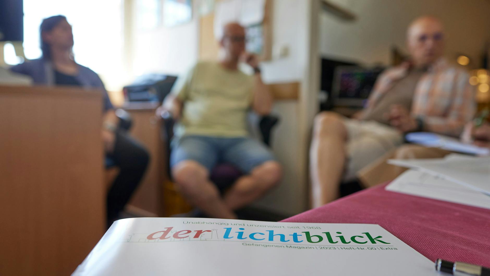 Eine Ausgabe der Gefangenenzeitung Der Lichtblick liegt auf einem Tisch im Zellentrakt der Justizvollzugsanstalt Tegel, während im Hintergrund drei Redaktionsmitglieder sitzen.