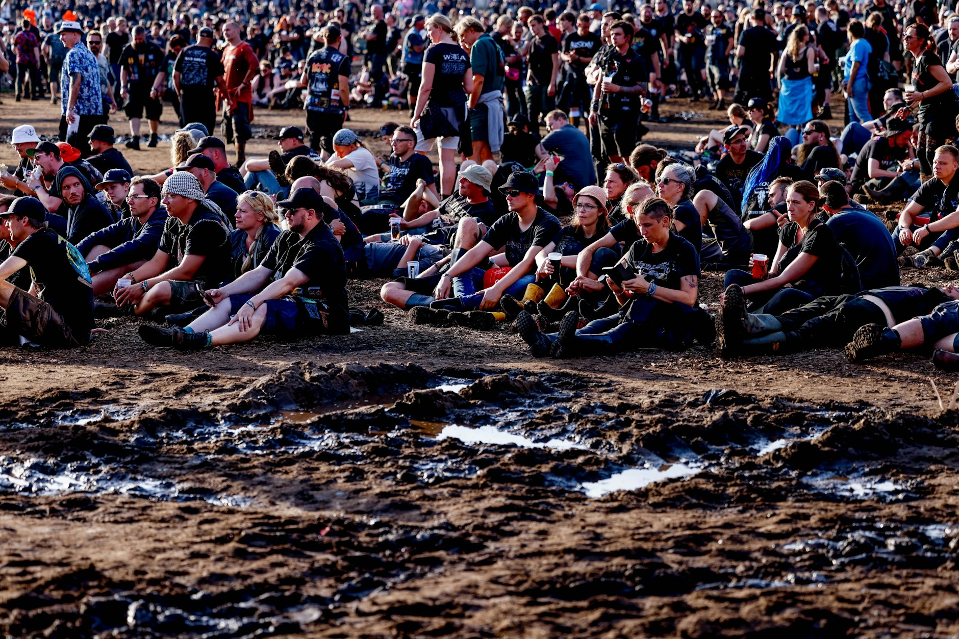 Schleswig-Holstein, Wacken: Festivalbesucher sitzen beim Wacken Open Air neben den abtrocknenden Schlammflächen auf dem Veranstaltungsgelände.