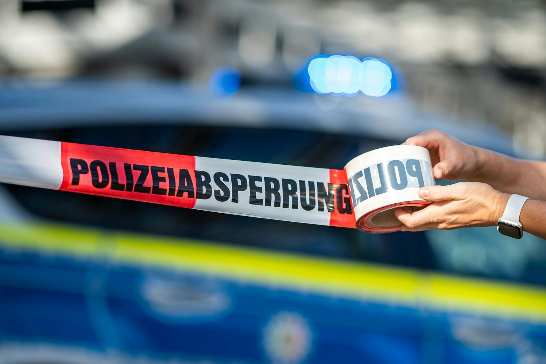 ILLUSTRATION - Ein Absperrband wird vor einem Polizeiwagen ausgerollt.
