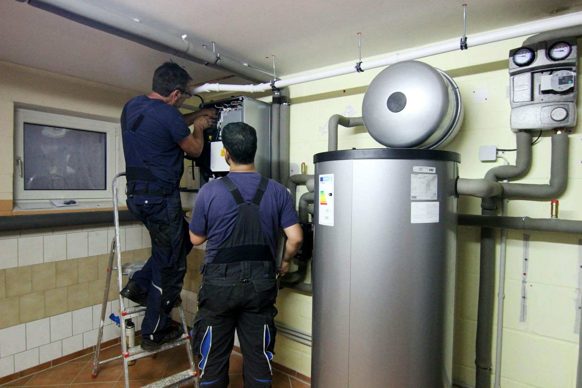 PRODUKTION - Zwei Handwerker stellen eine gerade installierte Wärmepumpe im Keller einer Doppelhaushälfte ein.