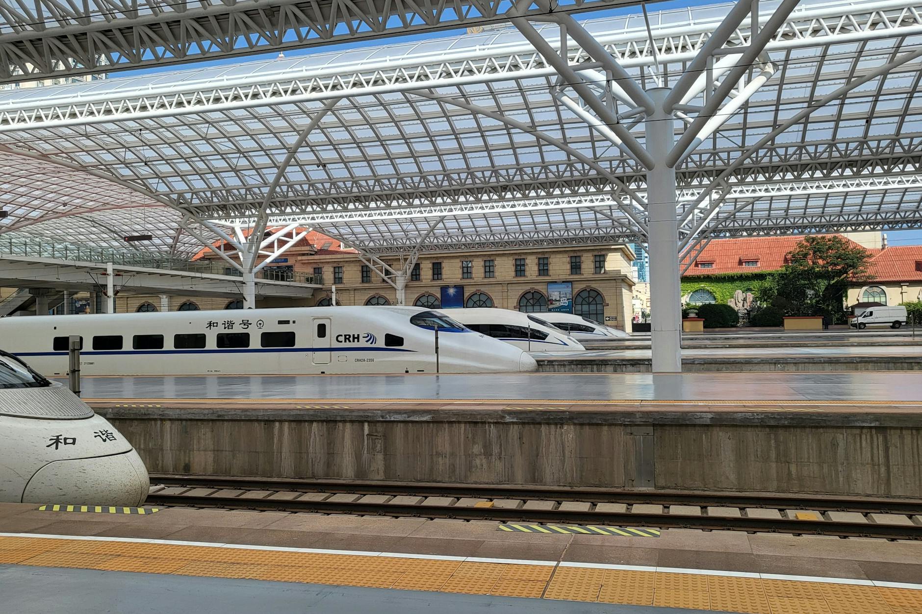 Bahnhof von Qingdao, vier Hochgeschwindigkeitszüge stehen bereit. Hinten: Teile des erhaltenen sanierten und erweiterten Bahnhofsgebäudes aus deutscher Kolonialzeit.