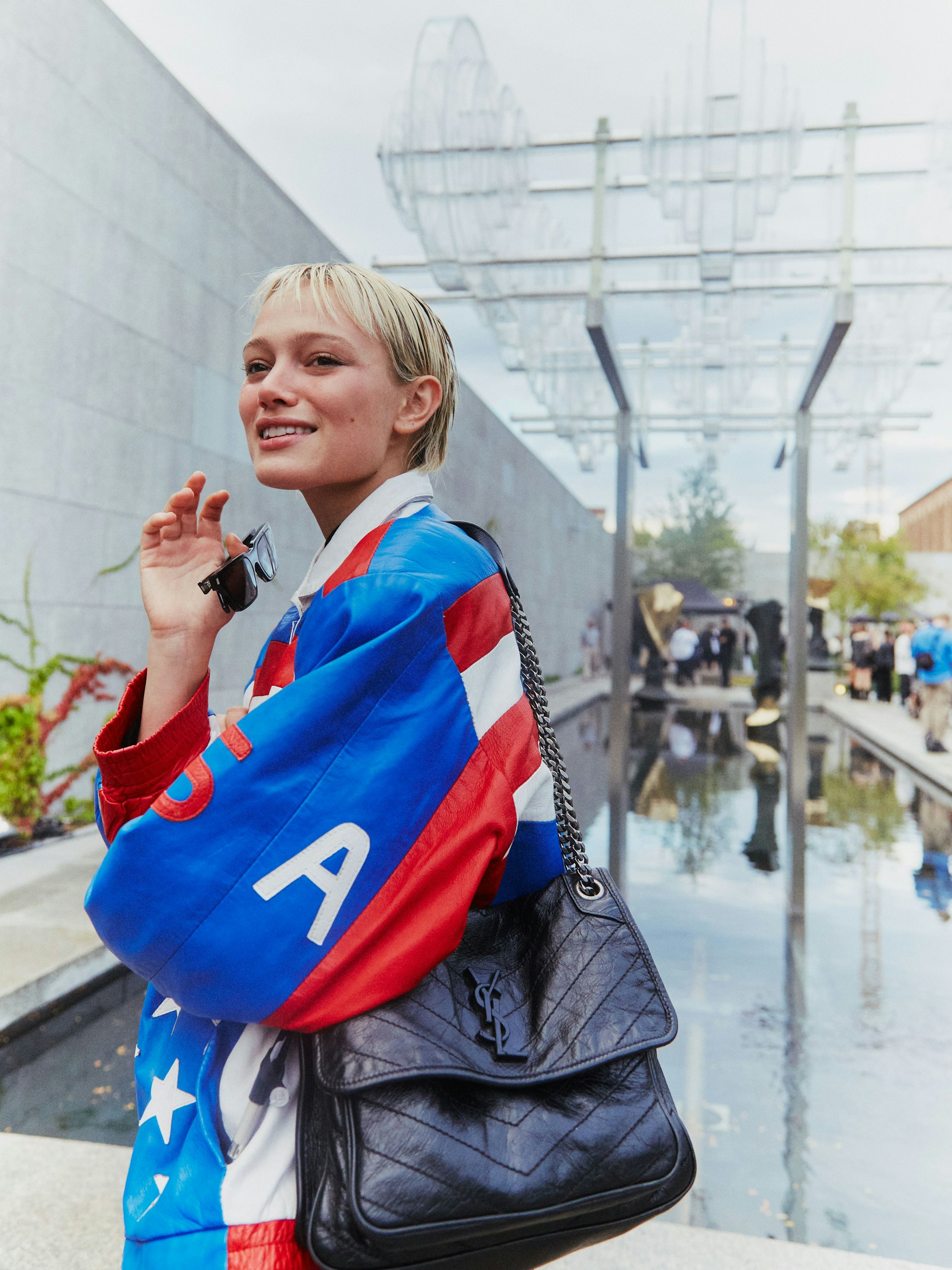 Zum USA-Look trägt Simona Tasche und Sonnenbrille von Saint Laurent. Die Sneaker sind von Nike. Im Hintergrund sieht man den BAW Garten, eine hochkarätig bespielte Festival-Location während der Berlin Art Week 2023.