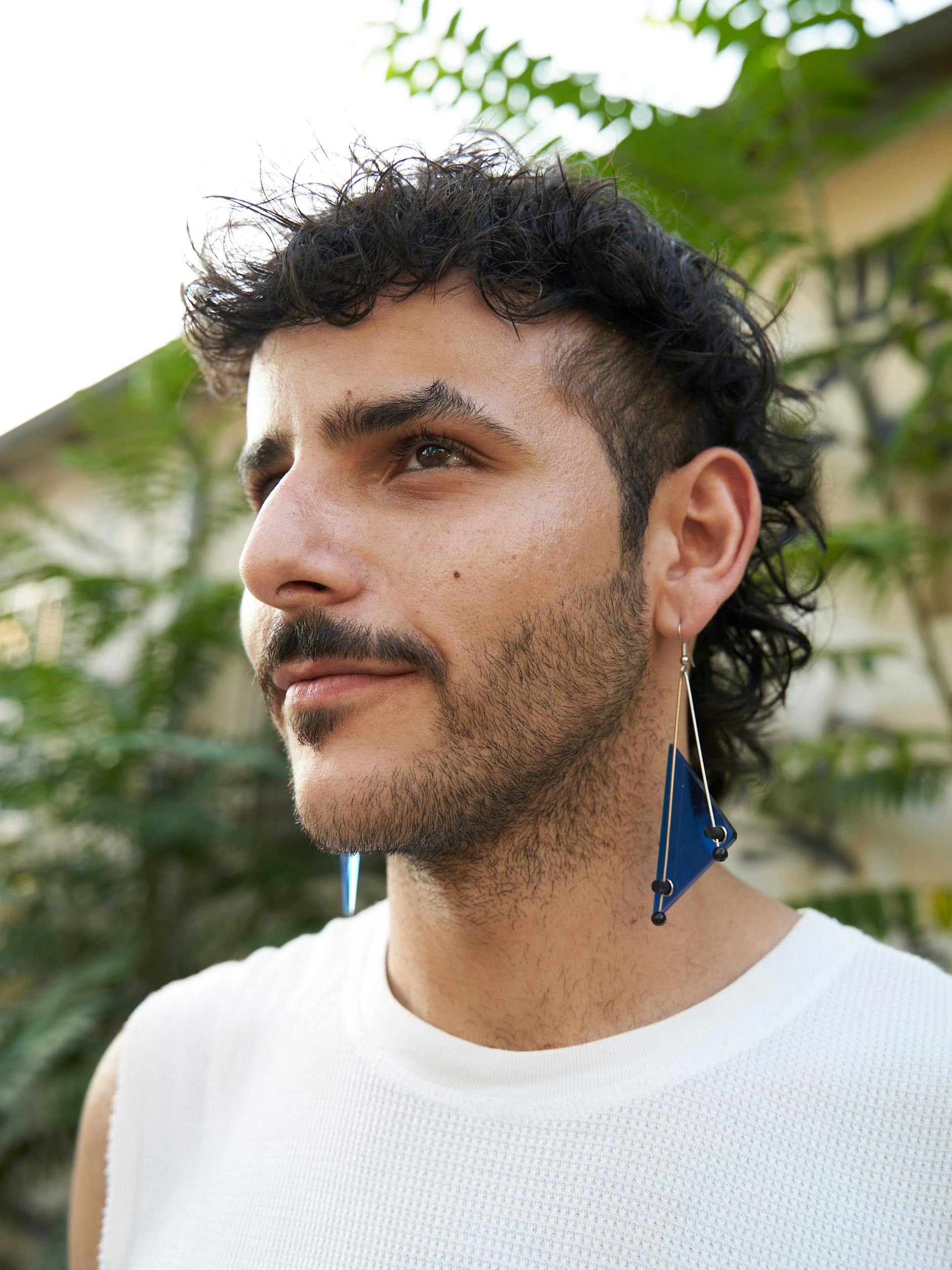 Mo trägt passend zur Hose blauweiße Ohrringe, die wie eine schmuckgewordene 80er-Jahre-Grafik wirken. Bezüglich Ohrschmuck sei er „obsessed“, sagt Mo. Sie seien ein Symbol für eine Freiheit, die er in seiner Heimat vermisst. In Syrien sei man sehr restriktiv bezüglich der Kleidung und des Schmucks. Als Mann könnte er diese Ohrringe dort nicht problemlos tragen.