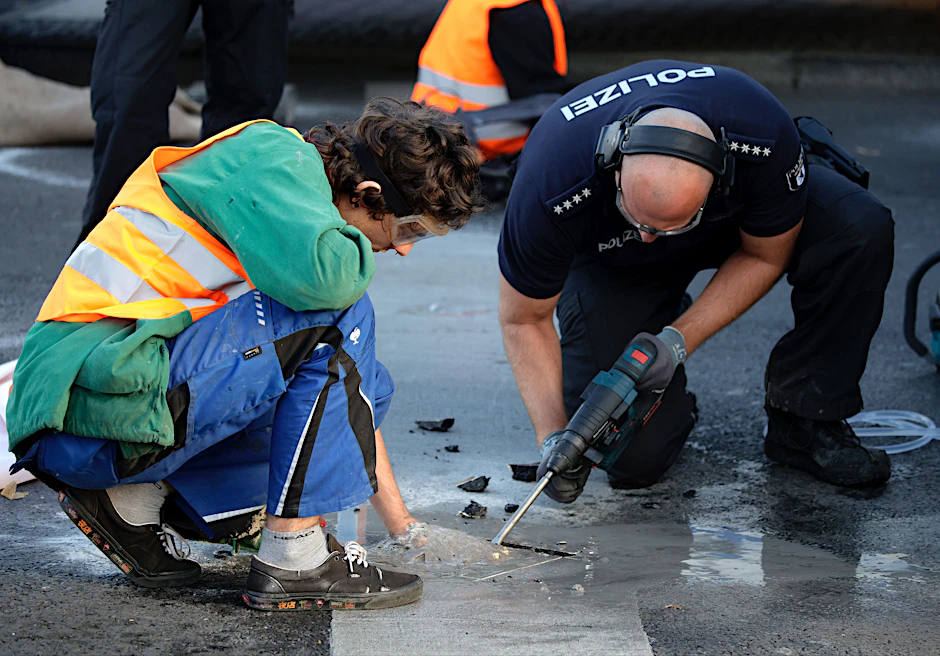 Berlin: Ein Polizist entfernt mit einem Bohrer einen Teil Asphalt, um einen Aktivisten von der Straße zu lösen. Am Dienstag fanden erneuet Protestaktionen der Klima-Aktivisten der Letzten Generation statt.
