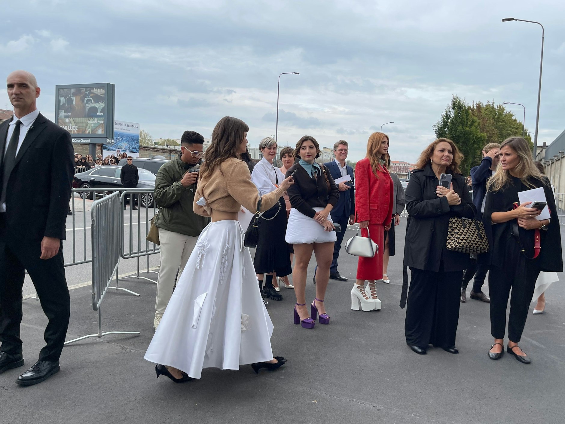 Eine Besucherin der Prada-Show trägt einen langen, weißen Rock mit floralen Applikationen aus der aktuellen Kollektion. Ihren Pulli hat sie keck zum Crop-Top gerafft, das kann man übrigens toll zu Hause nachmachen. Mittig im Bild trägt eine Dame den gepolsterten Mini von Prada, den es diesen Winter zu kaufen gibt. Da hat man das Sitzkissen stets mit dabei, praktisch.