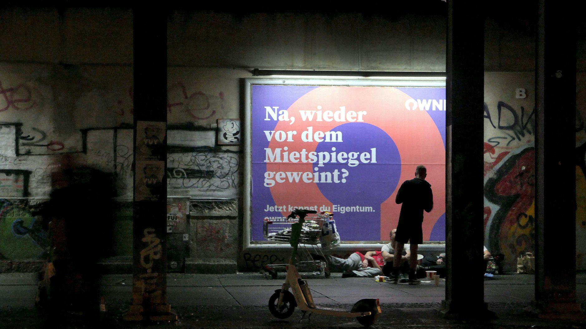 Obdachlose kampieren in Berlin vor einem Plakat, das für Wohneigentum wirbt.