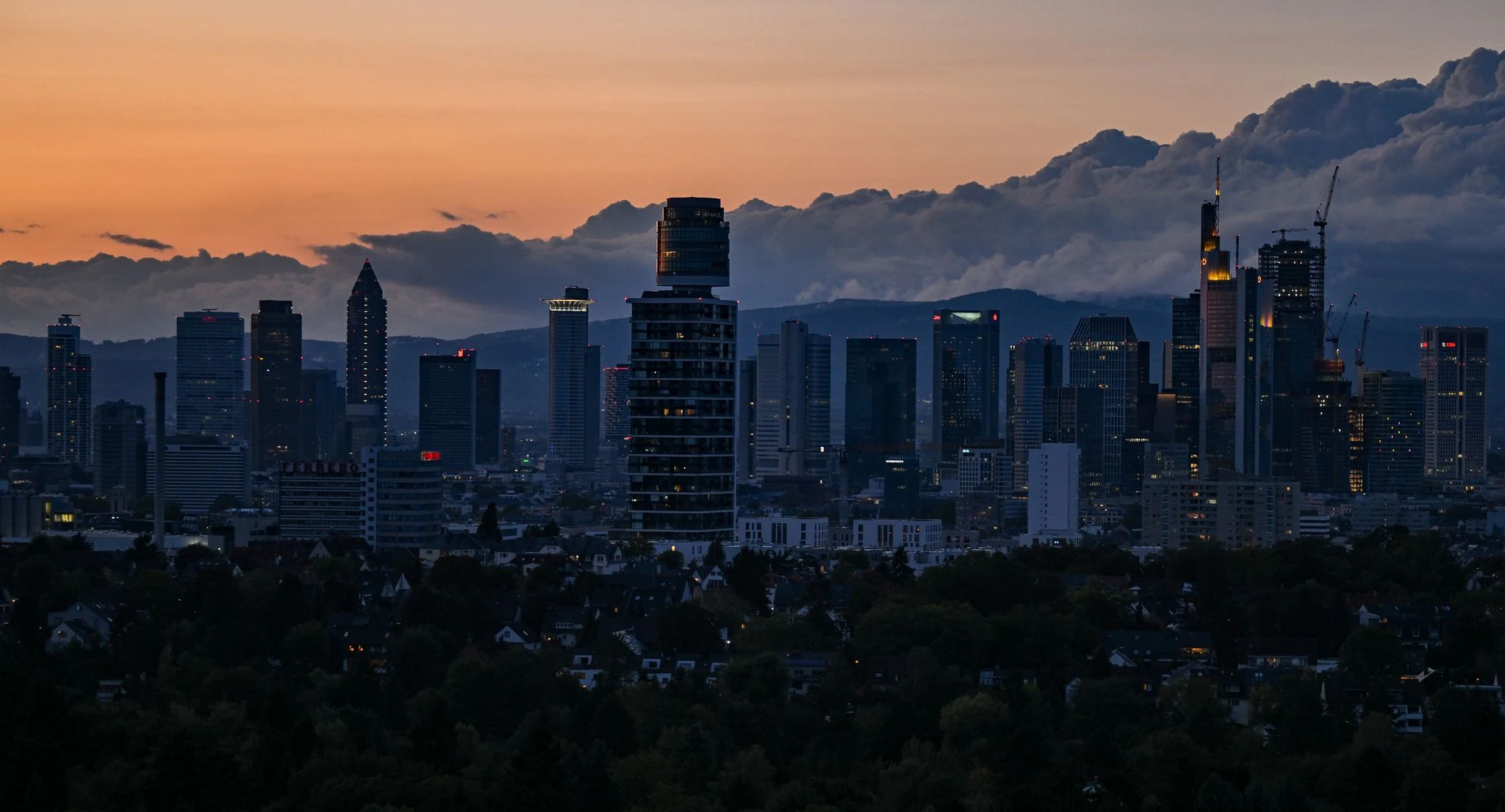 Während der Abendhimmel orange gefärbt ist und Wolken über die Taunushöhen ziehen, sind die Hochhäuser der Bankenstadt Frankfurt schwach beleuchtet.  