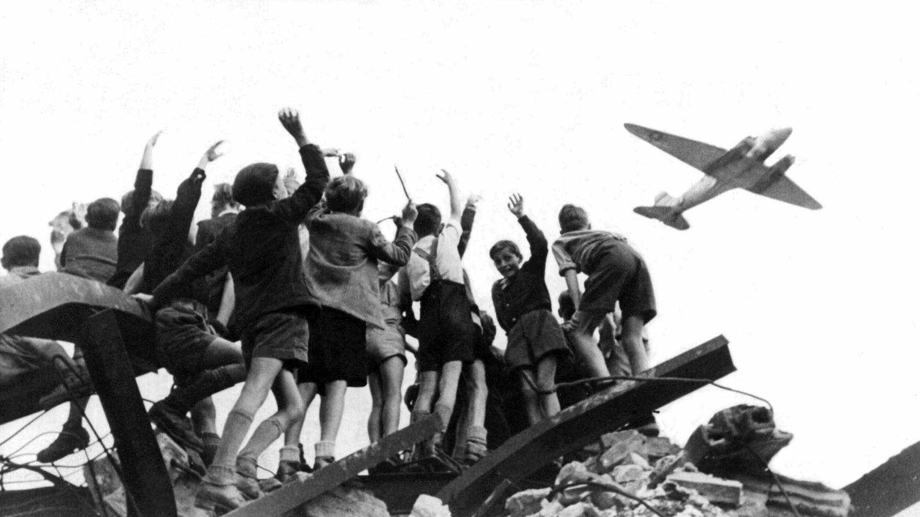 West-Berliner Jungen, die auf einem Trümmerberg stehen, begrüßen winkend ein amerikanisches Transportflugzeug, das Versorgungsgüter nach West-Berlin bringt.