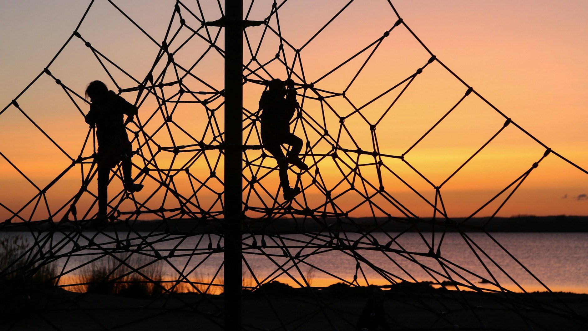 Kinder klettern vor der untergehenden Sonne auf einem Klettergerüst am Strand. Sich draußen austoben – das können nicht alle Kinder direkt vor ihrer Tür. Doch manchmal scheitert es auch an den Eltern. Sie meinen es gut, bewirken aber das Gegenteil.