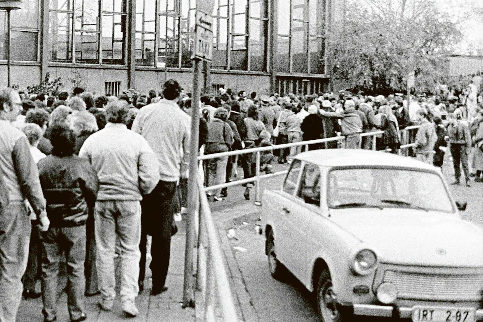 Am 10. November 1989 steht eine Menschenmenge dichtgedrängt vor dem Tränenpalast am Bahnhof Friedrichstraße und will in den Westteil Berlins. Im Halteverbot steht ein Trabi, der nun Fragen aufwirft.