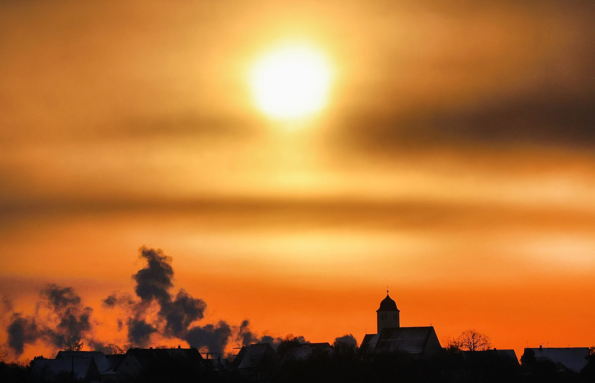 Winterlicher Morgen: Rauch dringt bei Minusgraden in Oberstetten auf der Schwäbischen Alb aus Kaminen. Im Hintergrund geht die Sonne hinter Wolken auf.  