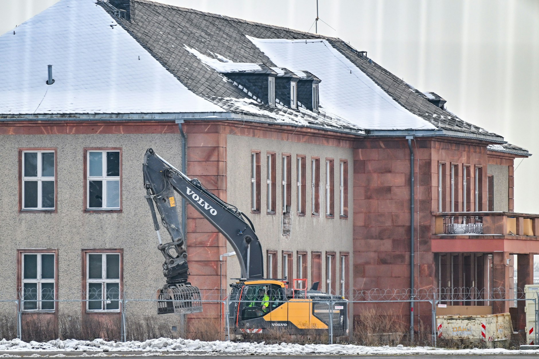 Blick durch einen Zaun auf einen Bagger vor dem zum Abriss vorgesehenen Generalshotel auf dem ehemaligen Flugplatz Schönefeld. 