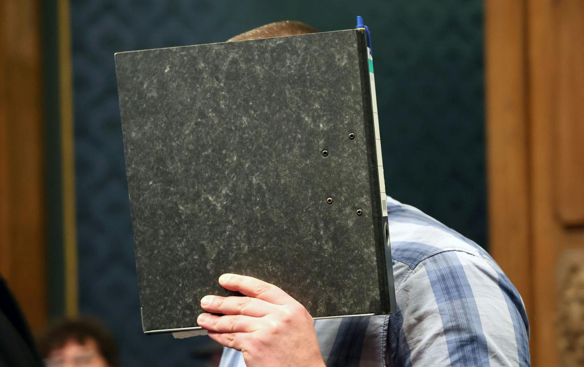 Der angeklagte Feuerwehrmann mit einem Aktendeckel vor dem Gesicht im Gerichtssaal.