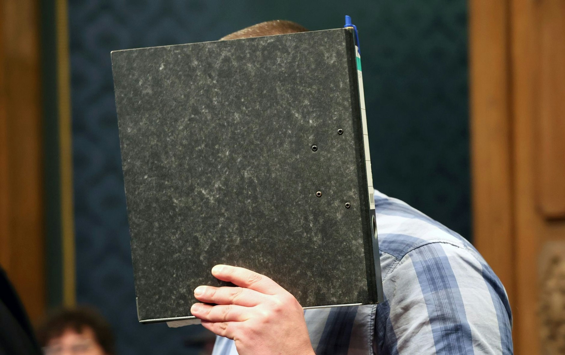 Der angeklagte Feuerwehrmann mit einem Aktendeckel vor dem Gesicht im Gerichtssaal.  